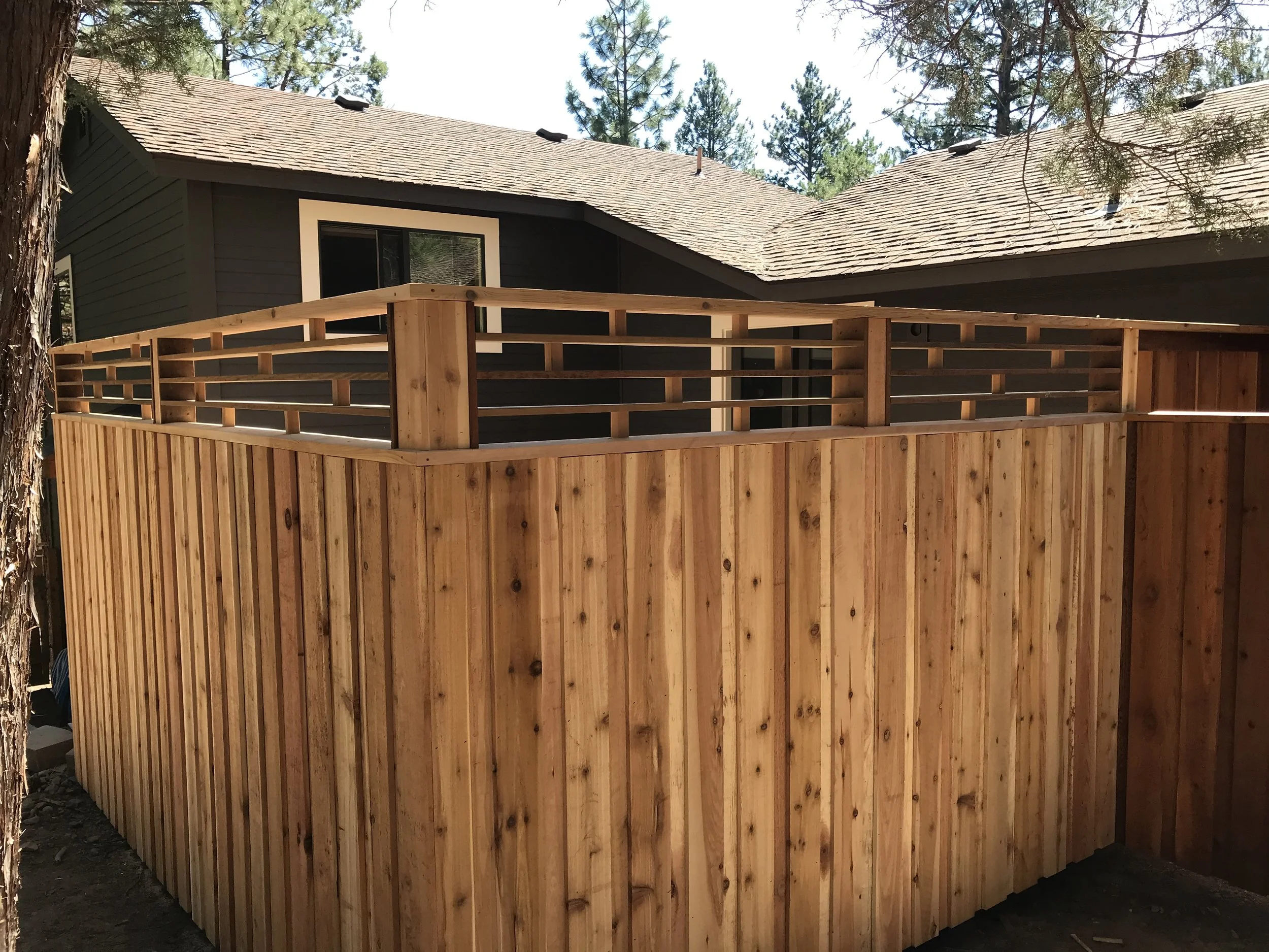 Wooden deck under construction with vertical planks and a railing, adjacent to a house with a gray exterior and shingled roof, surrounded by trees.
