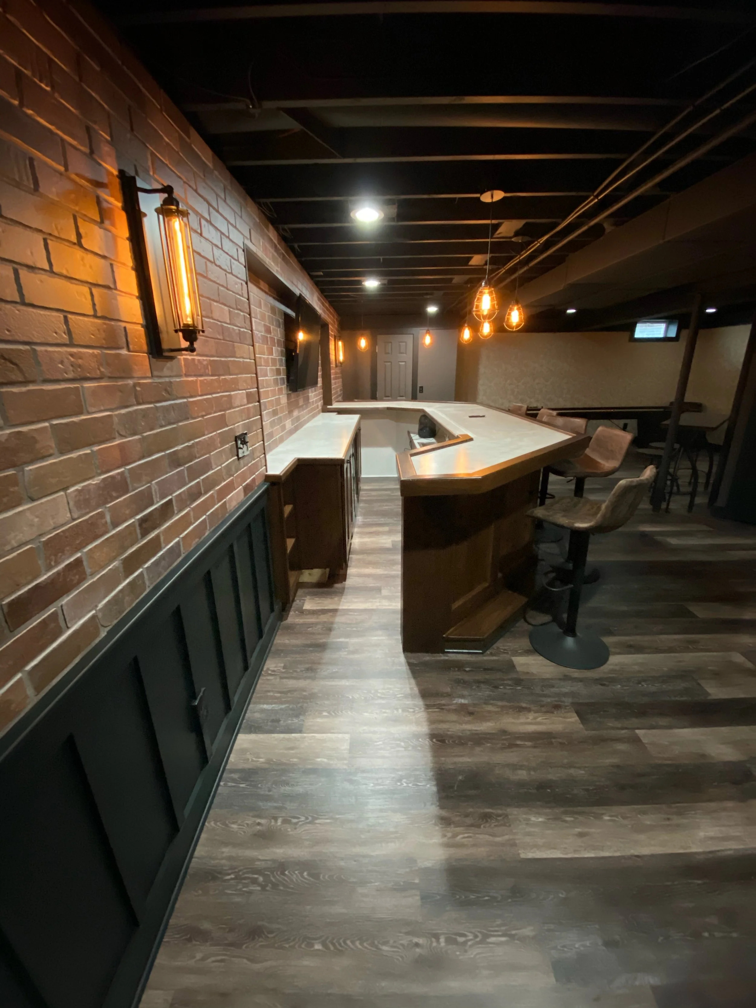 Interior of a bar with a brick wall, wooden bar counter, bar stools, and warm hanging lights.