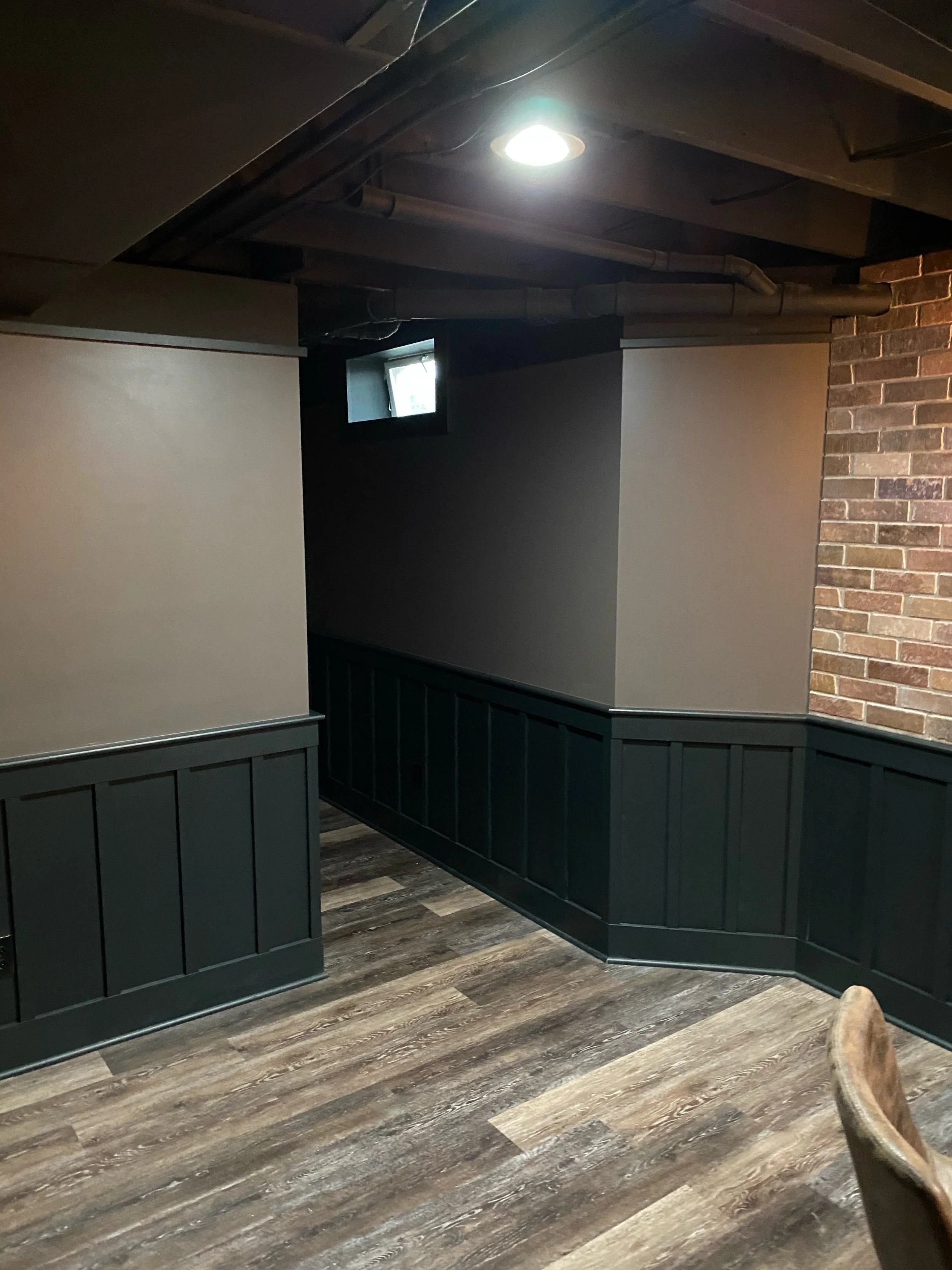 A corner of a room with a small window, dark walls, a brick wall, and wood flooring.