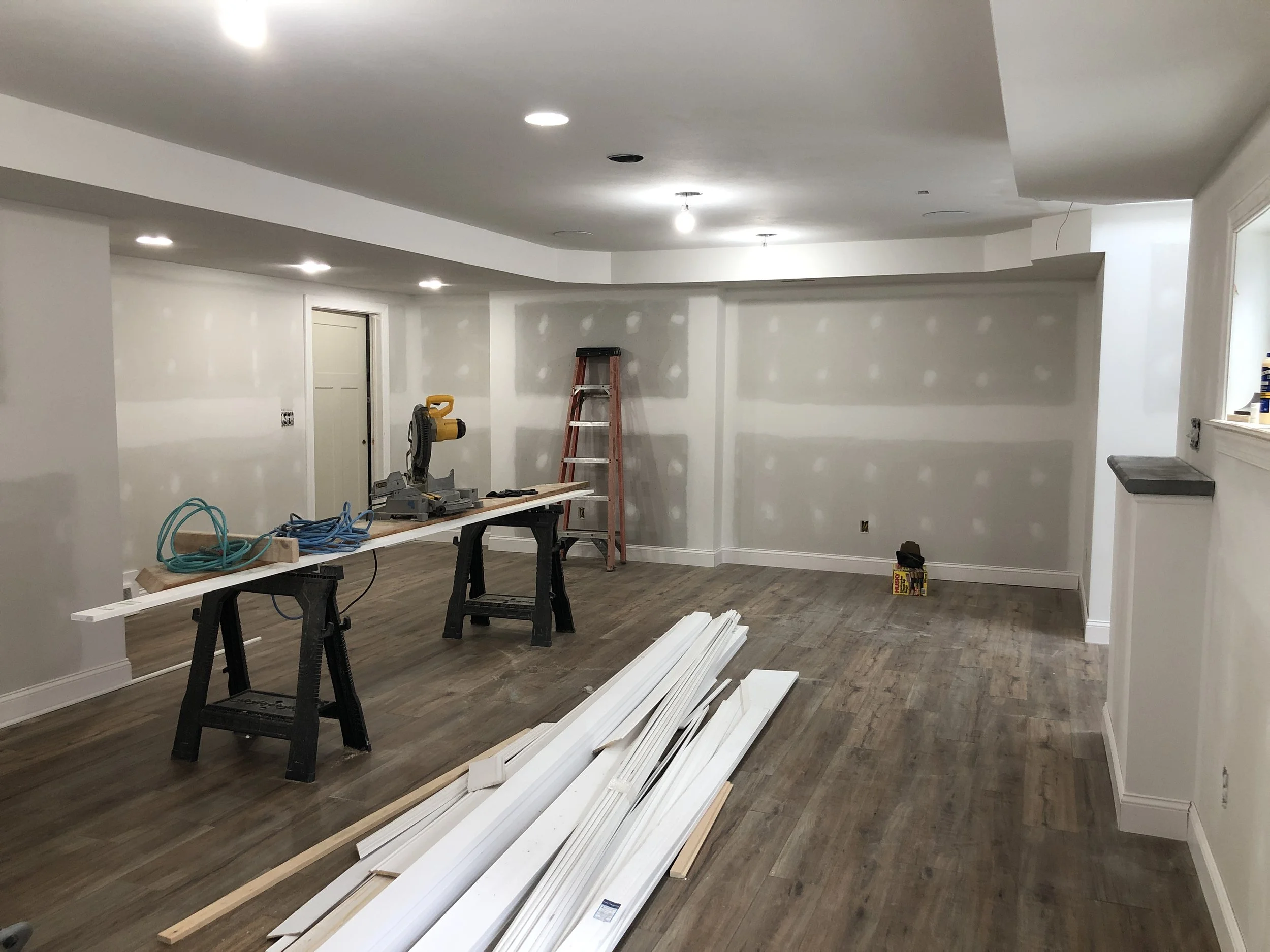Interior view of a room under renovation with drywall installation in progress, featuring a work table, ladder, construction tools, and flooring.