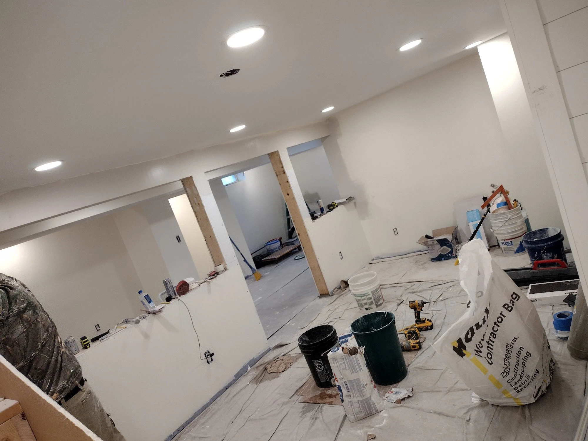 Construction site indoors with partially finished walls, construction tools, buckets, and materials scattered on the floor covered with plastic sheeting.