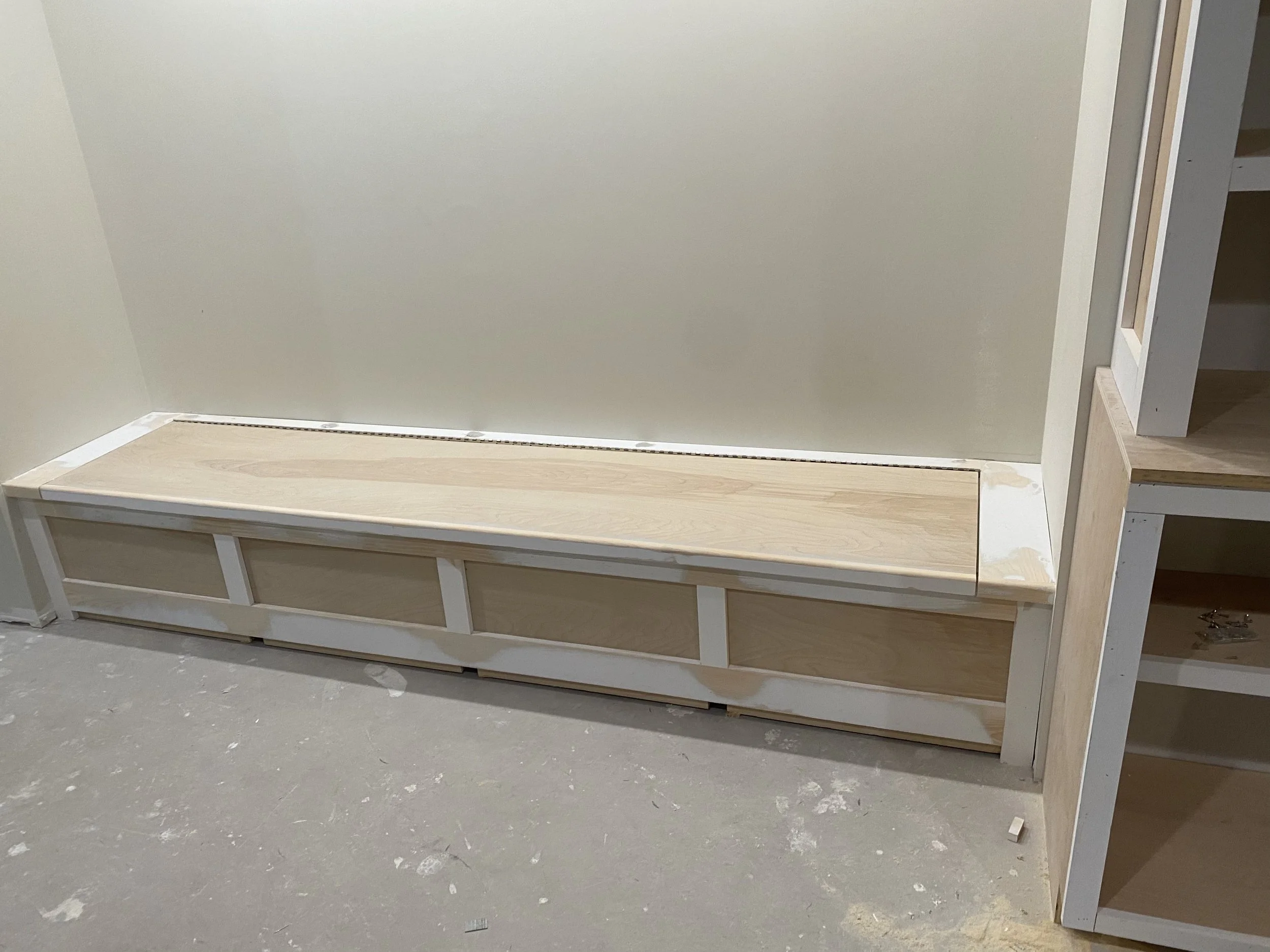 Wooden built-in storage bench with white trim in a room under construction, with drywall and unfinished flooring.