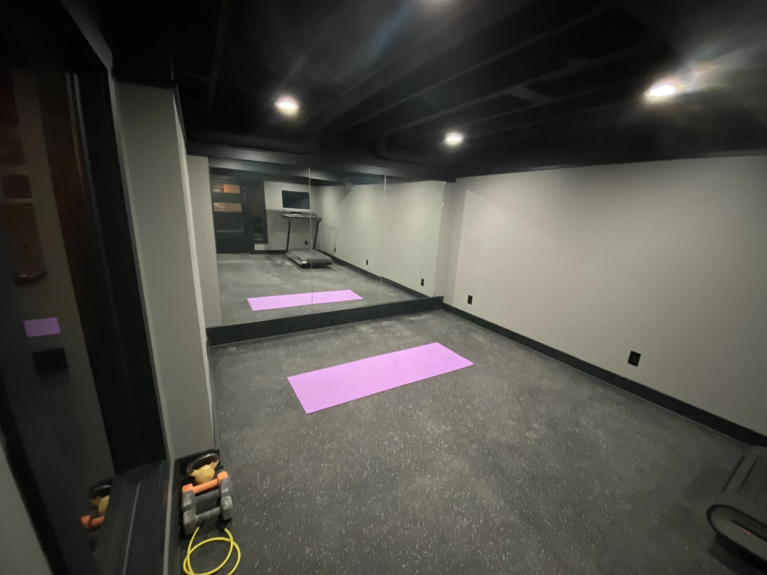 Empty home gym with two purple yoga mats on black rubber flooring, a treadmill, and a large mirror covering the back wall, with black ceiling and walls.