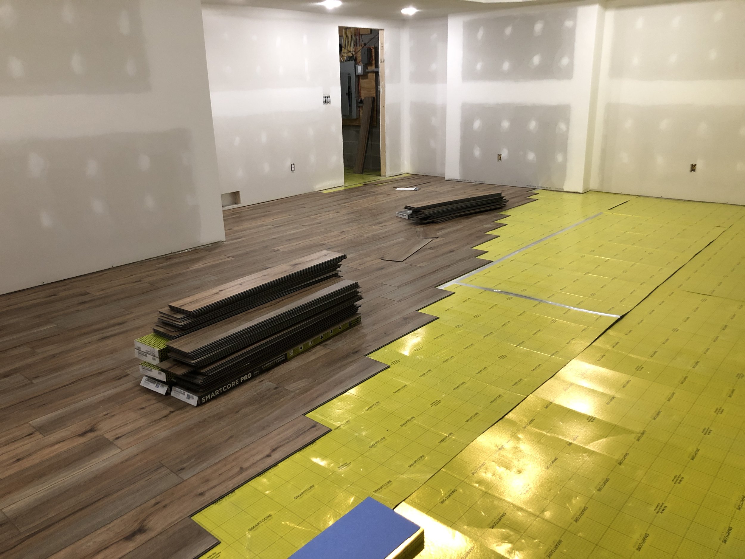 Room under construction with partially installed wooden flooring and yellow underlayment, construction materials stacked on the floor, and unpainted drywall walls.