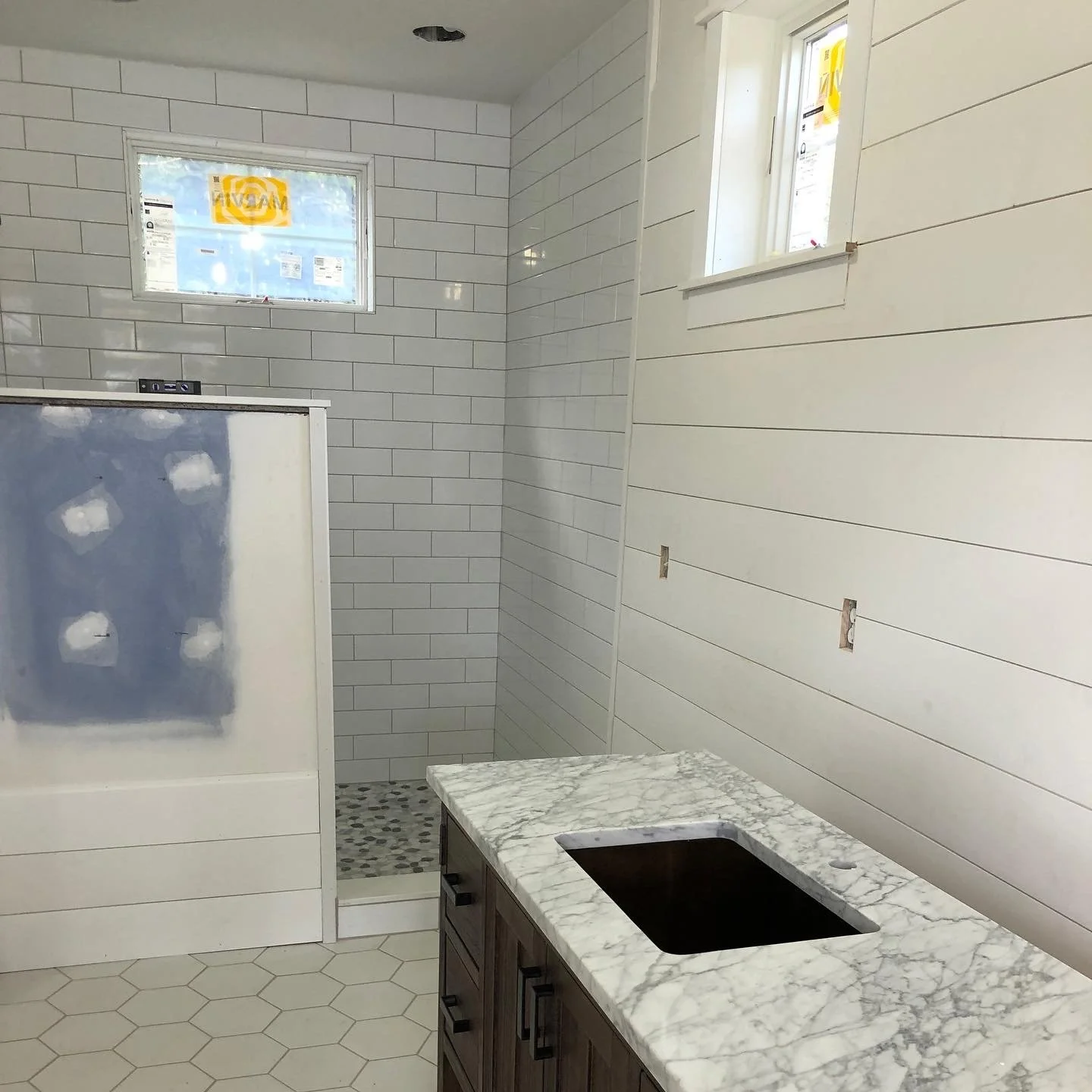 Bathroom with white shiplap walls, a small window, a marble countertop with an undermount black sink, and an adjacent shower area with white subway tiles and hexagonal floor tiles, under construction.