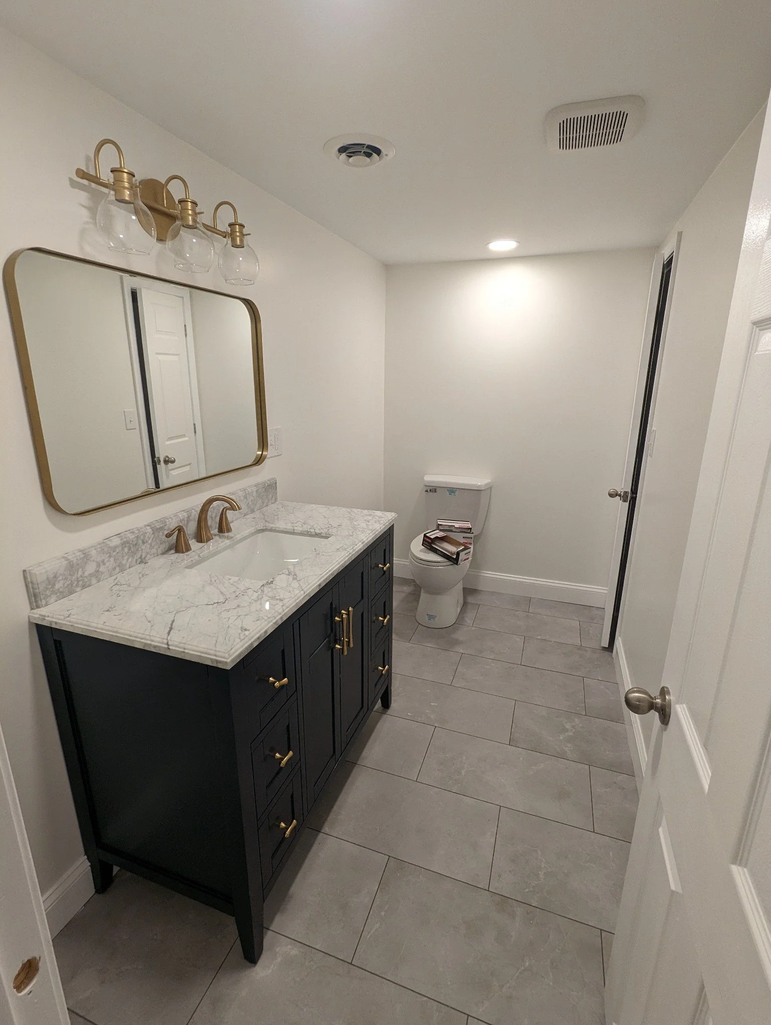 Modern bathroom with a navy vanity, white marble countertop, gold hardware, mirror, wall-mounted light fixtures, toilet with books on it, and gray floor tiles.