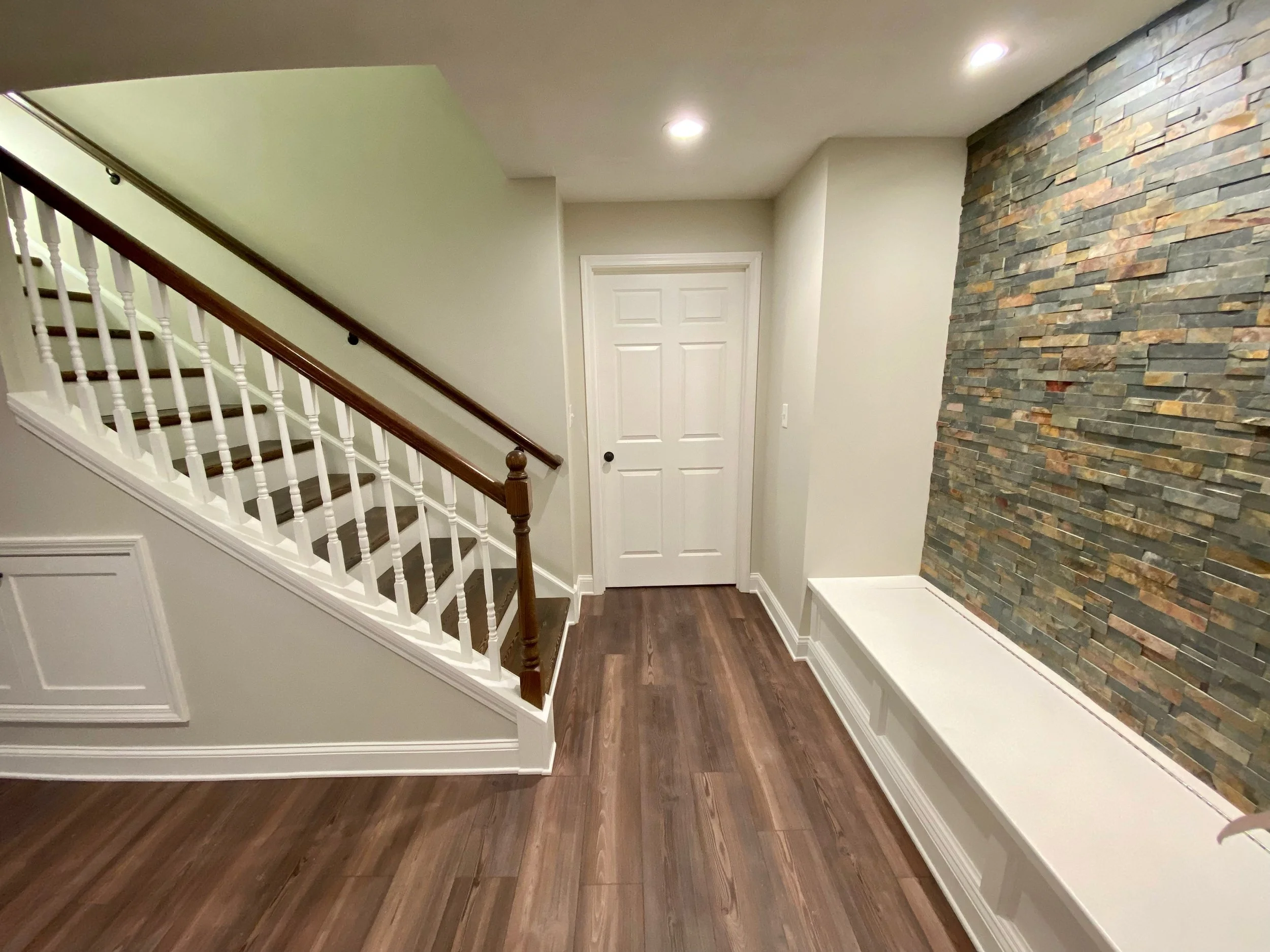 Interior view of a hallway with a staircase on the left, a white door at the end, and a textured stone accent wall on the right, with hardwood flooring and recessed ceiling lights.