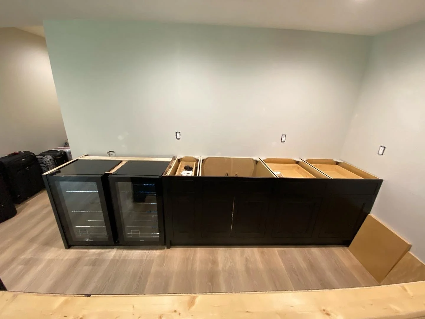Kitchen cabinet installation in progress with two glass-fronted wine coolers, and uninstalled cabinet sections along a wall with electrical outlets, and some luggage in the corner.