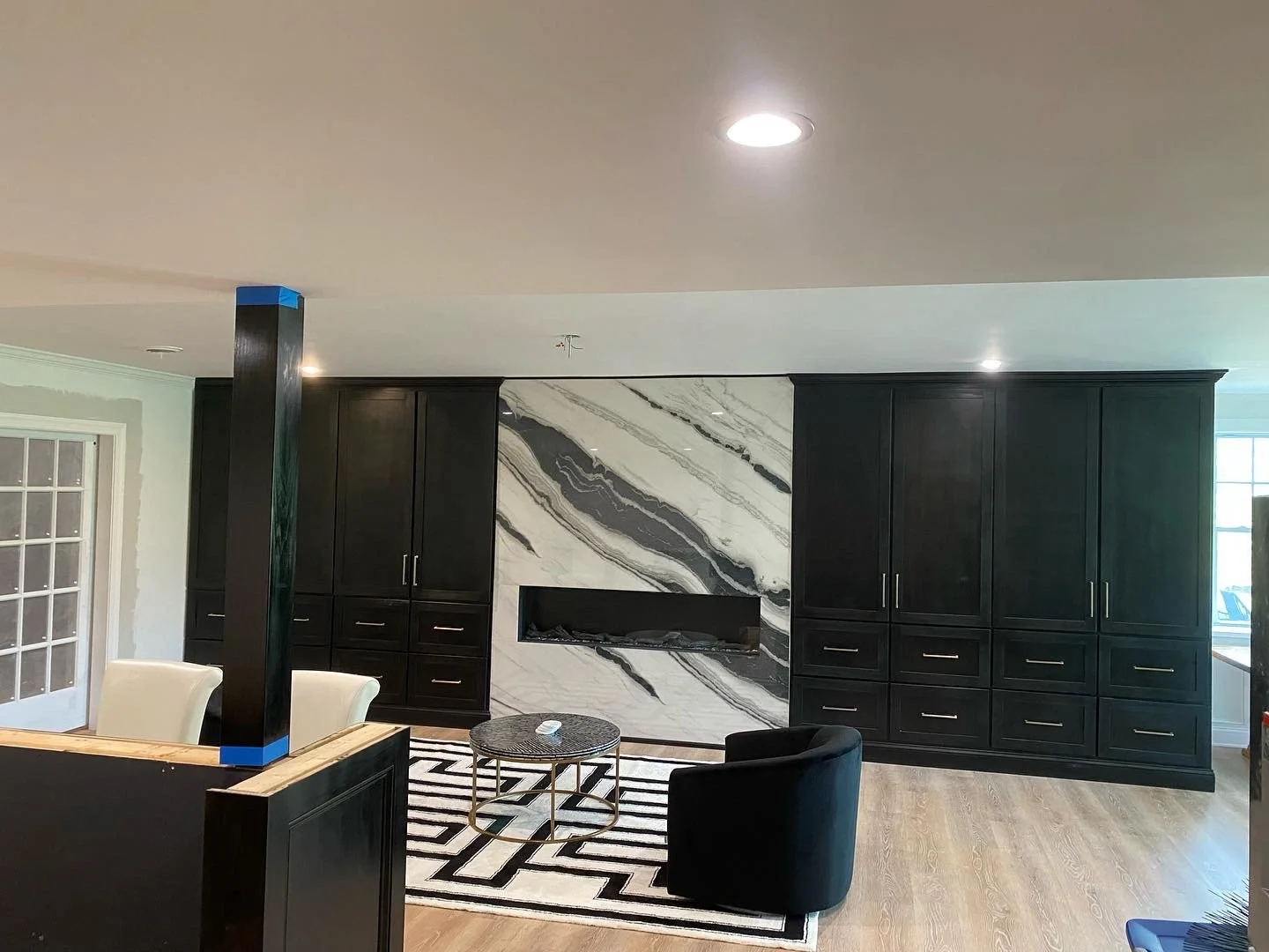 Living room with black cabinetry, marble wall feature with built-in fireplace, beige chairs, black armchair, round coffee table, patterned rug, and large window on the right.