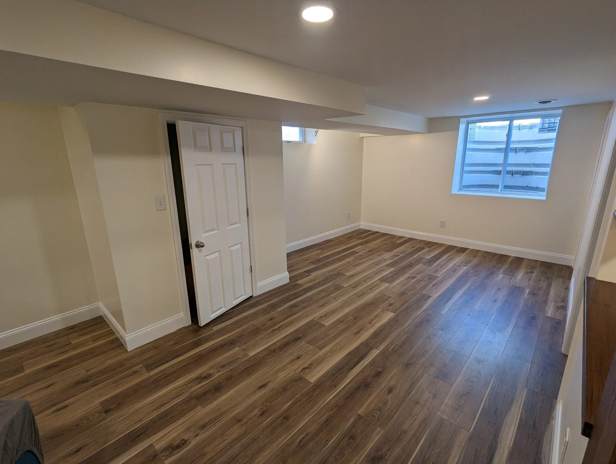 Empty room with wood flooring, white walls, a window with view of a neighboring building, and a closed door.