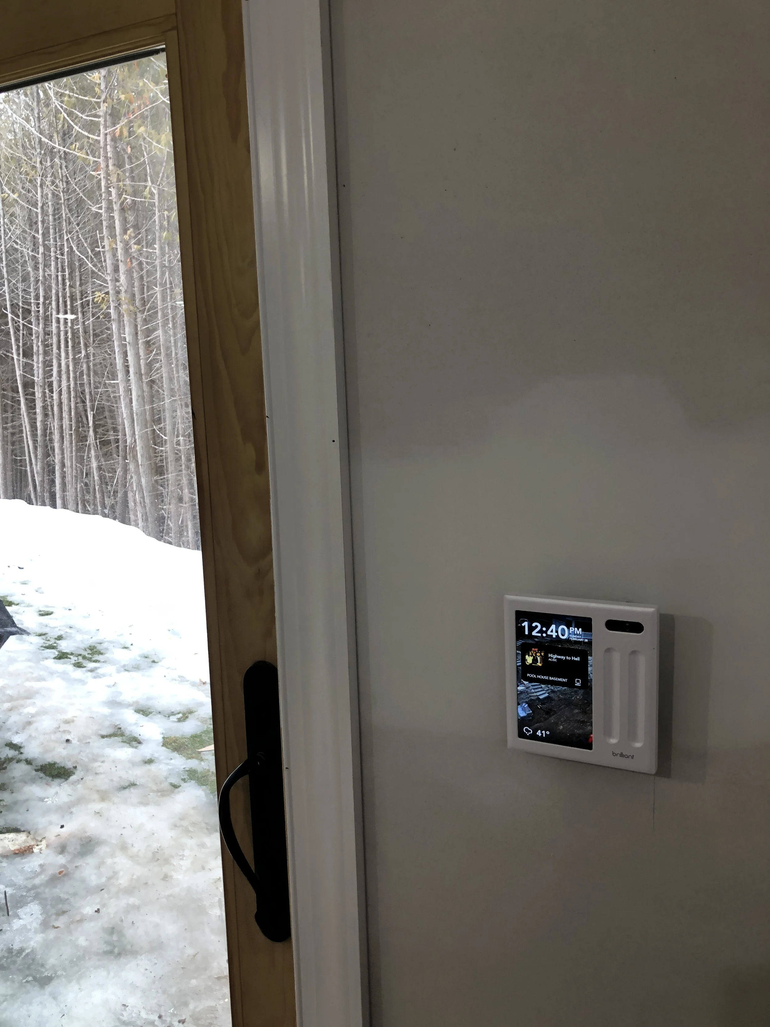 A smart home control panel mounted on a white wall inside a house, with a door and snow-covered outdoor area with trees visible in the background.