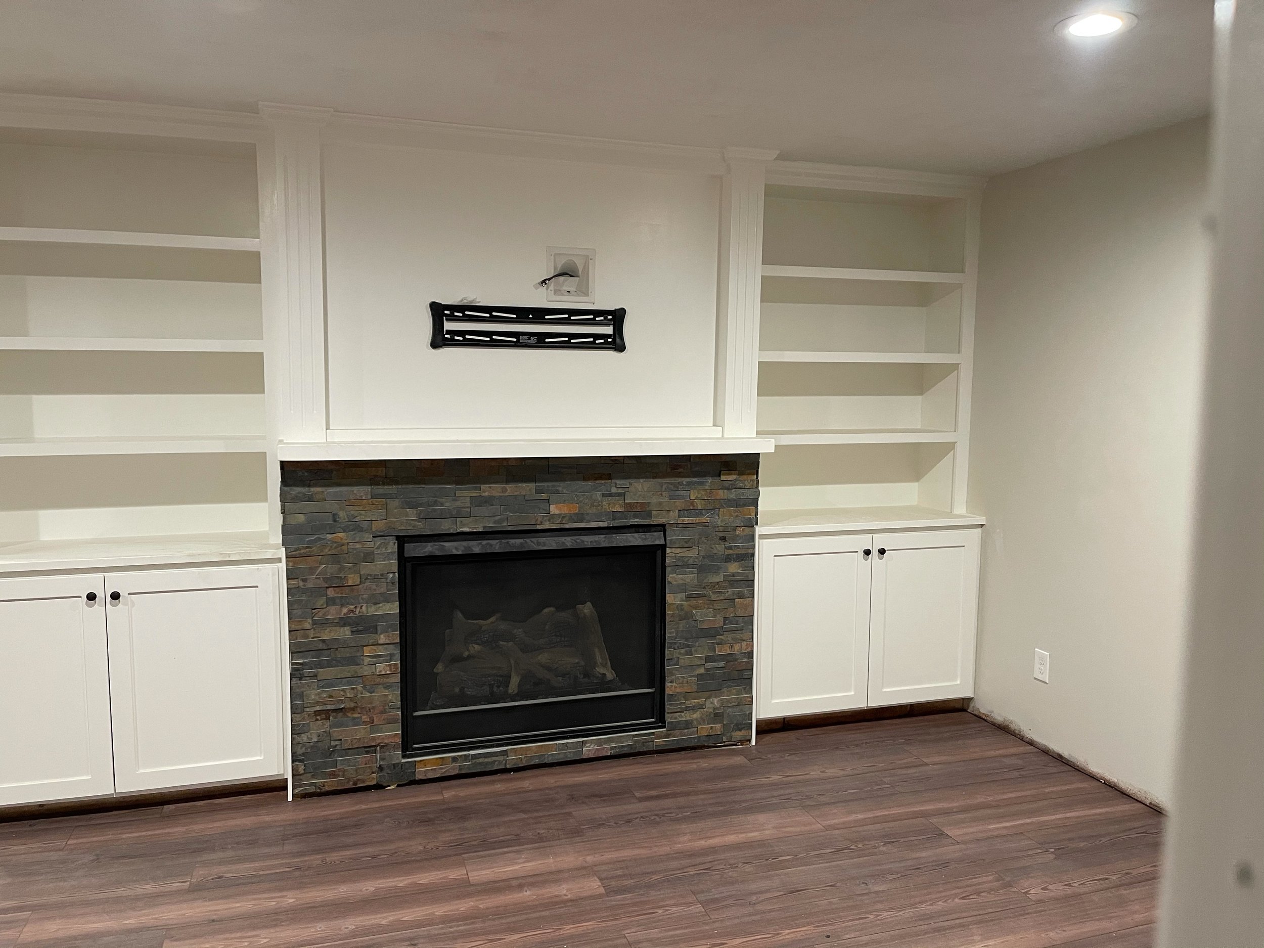 Living room with a wood-burning fireplace surrounded by stacked stone, built-in white bookshelves on either side, and a wooden floor.