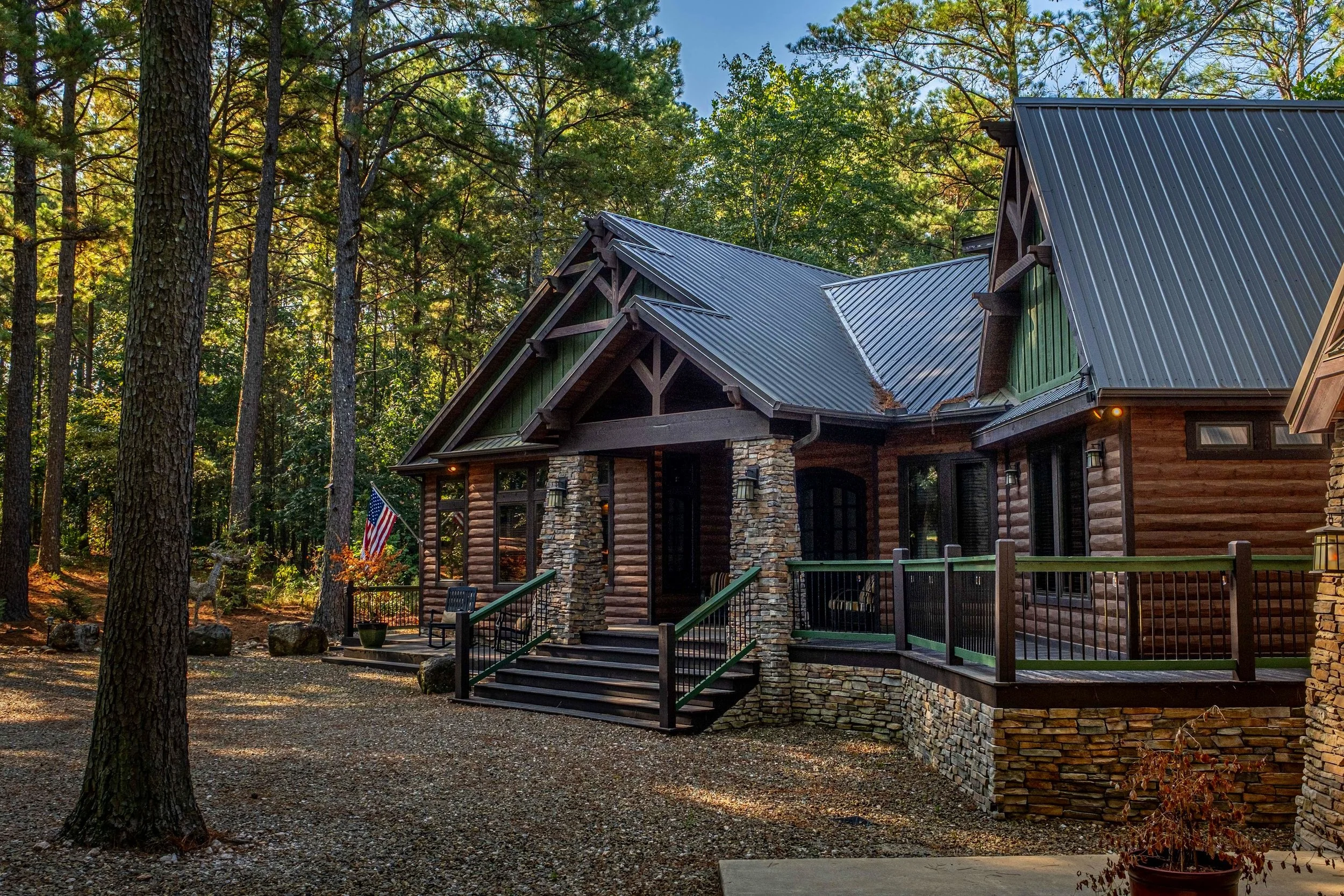 A rustic log cabin with stone pillars, green accents, and a metal roof, surrounded by tall pine trees in a wooded setting.