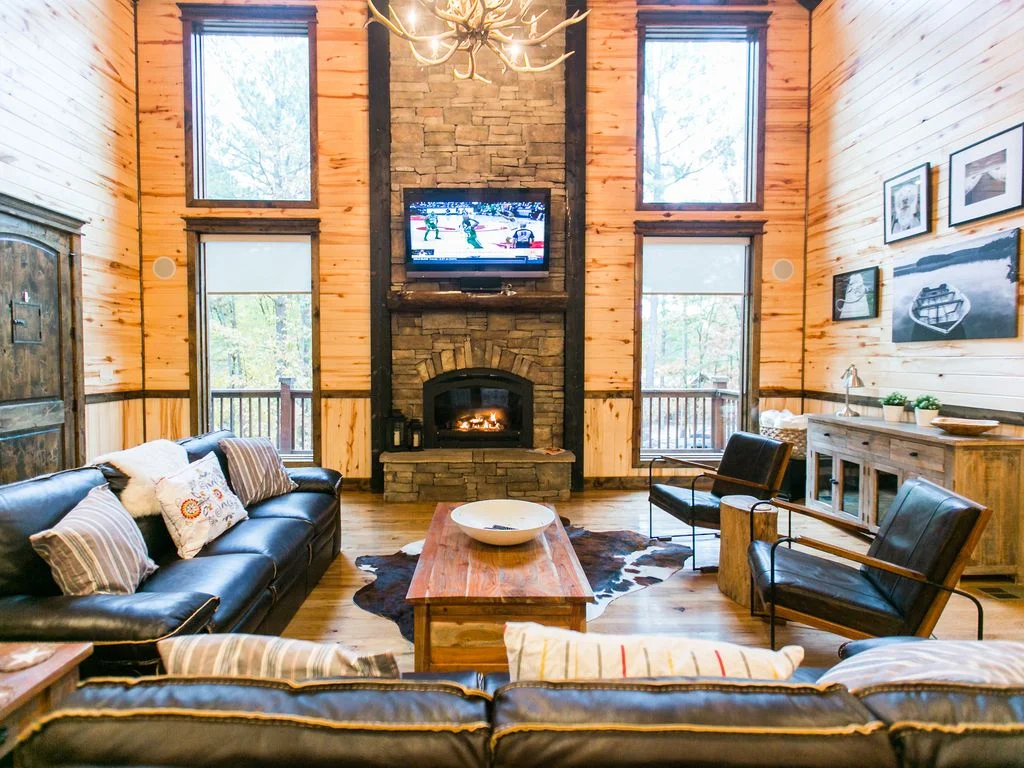 A cozy living room with wood-paneled walls, a stone fireplace with a TV mounted above, two large windows, a leather sofa with throw pillows, and two black armchairs around a wooden coffee table. There is a cowhide rug on the wooden floor and wall art