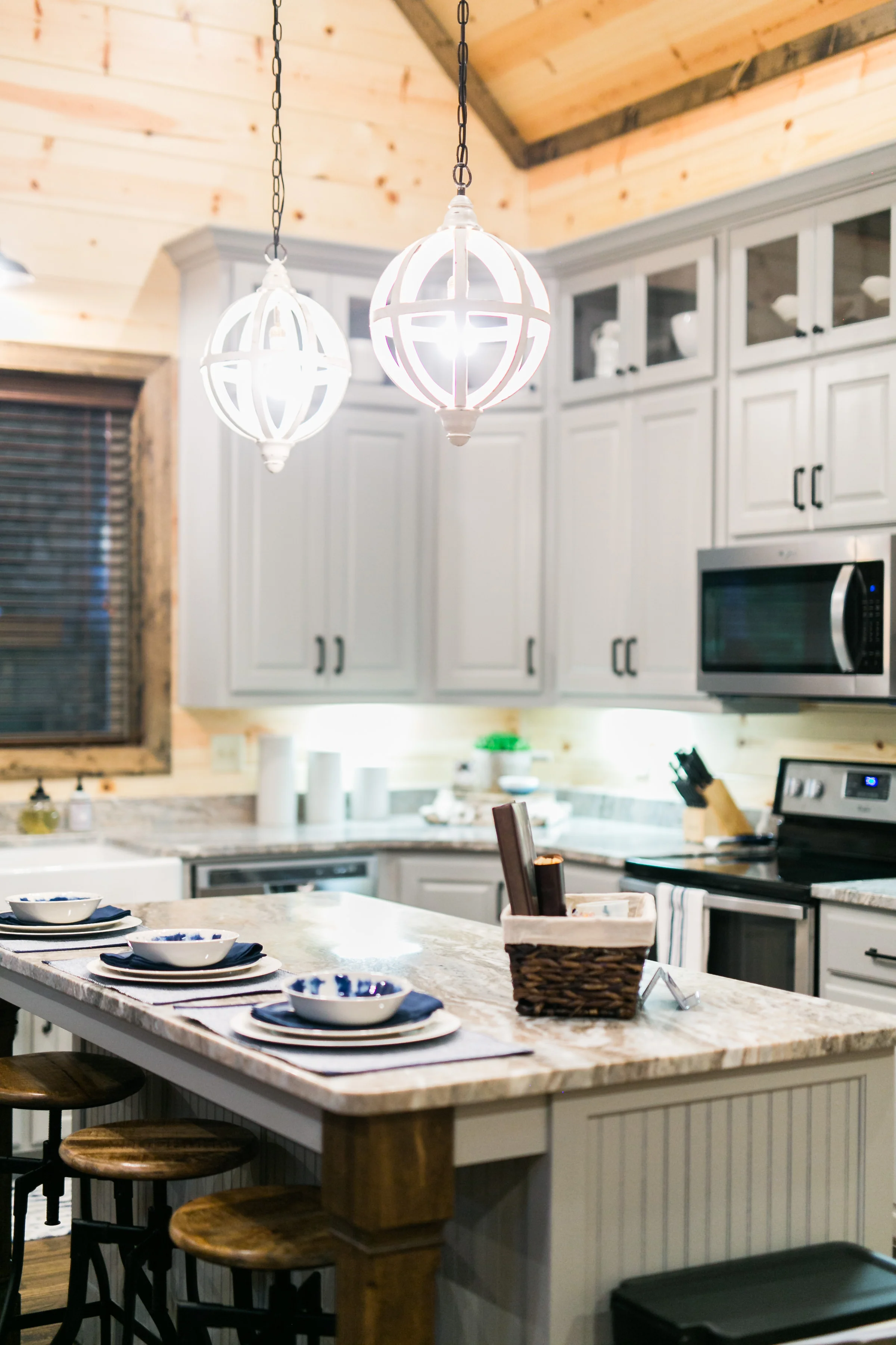 A cozy kitchen with light gray cabinets, a granite countertop, wooden accents, and two white pendant lights hanging above a kitchen island set with plates, bowls, and napkins.