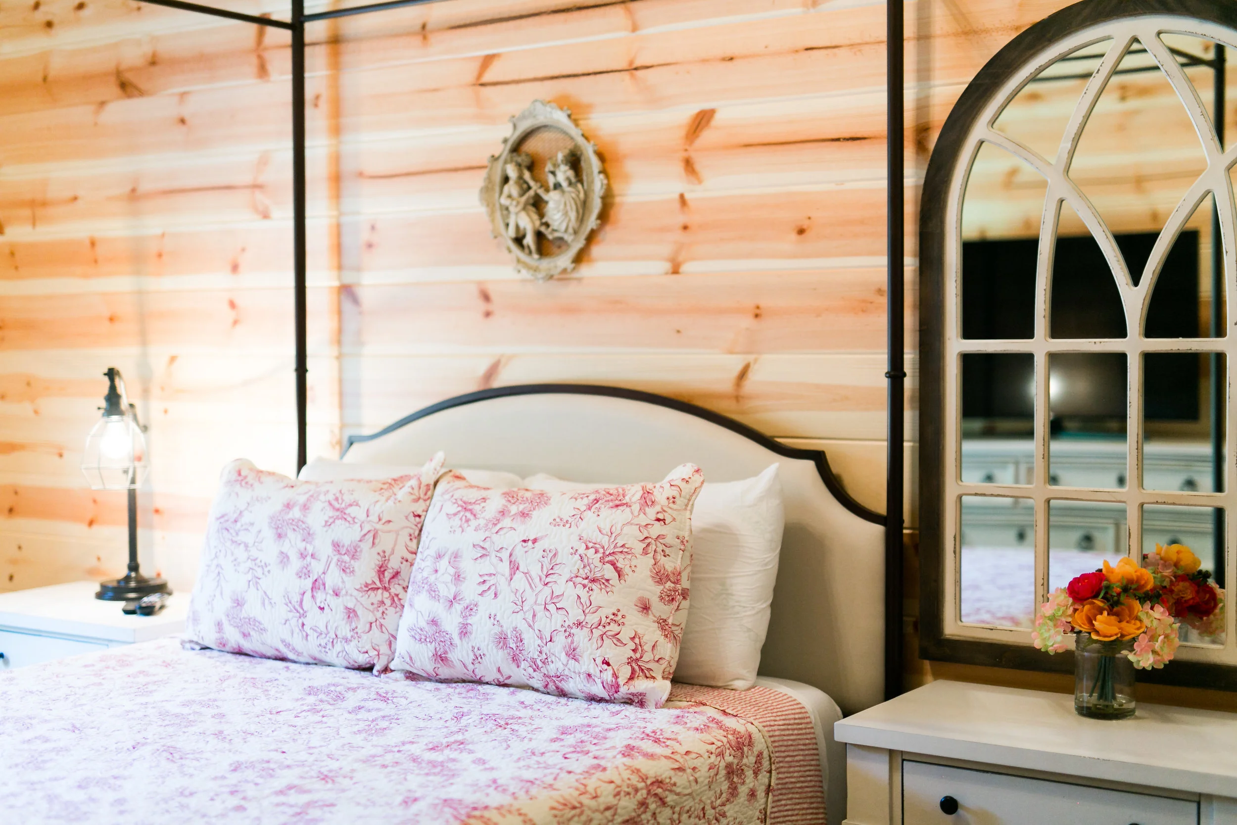 A bedroom with a wooden wall, a bed with white and pink floral bedding and pillows, a vintage mirror, a nightstand with a flower vase, and a wall art of cherubs.