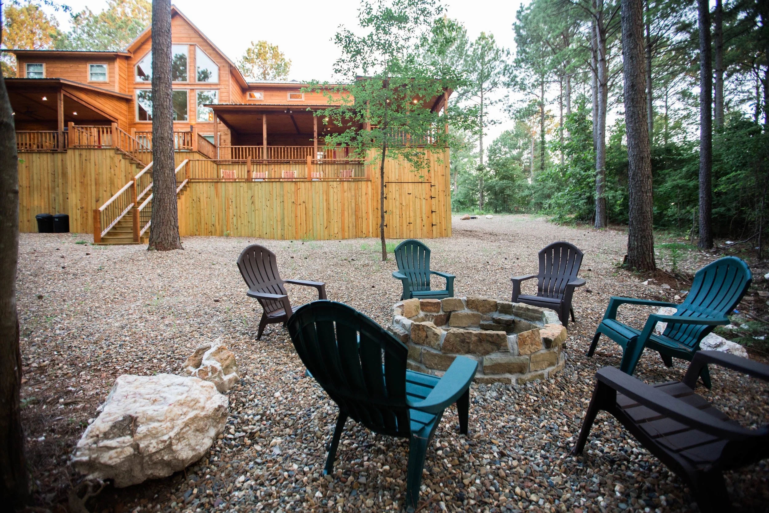 Outdoor backyard with a stone fire pit surrounded by six plastic chairs, tall trees, and a wooden house with a deck in the background.