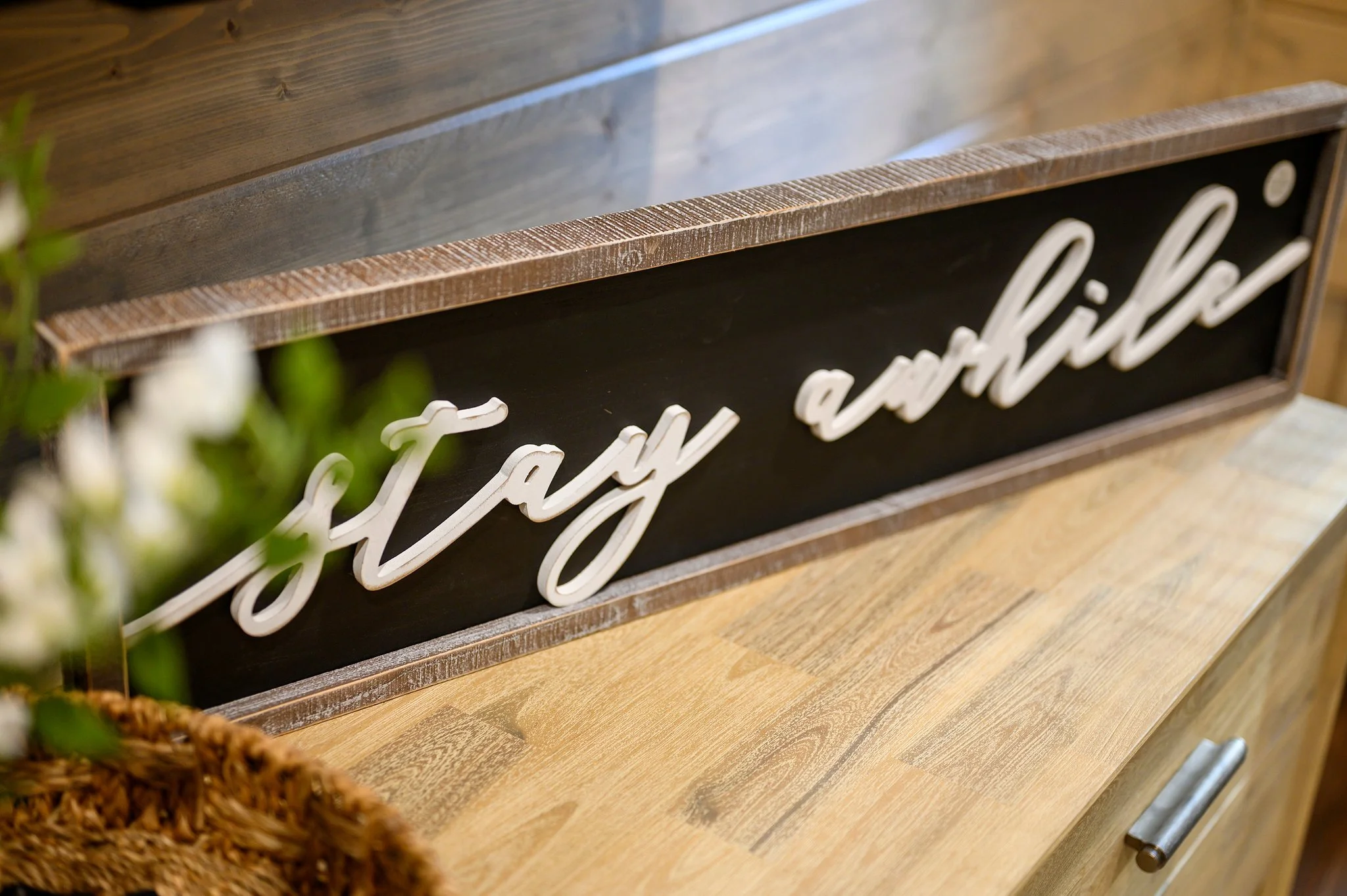 Decorative sign with the phrase 'stay awhile' in white cursive letters on a black background, surrounded by a wooden frame and placed on a wooden surface.