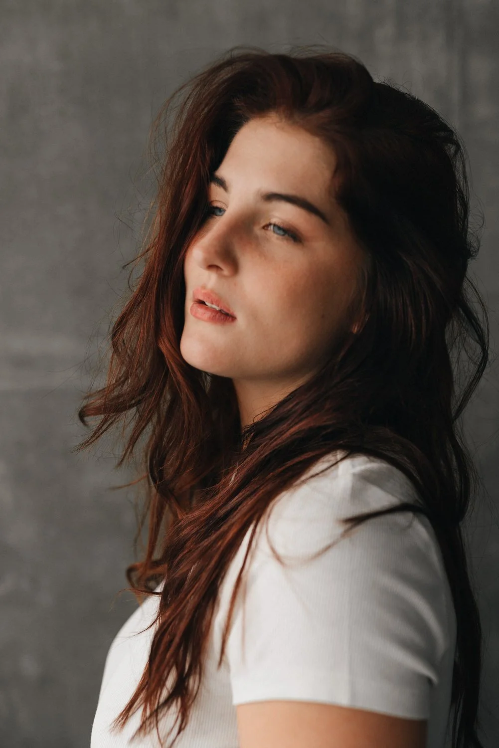 Nahaufnahme einer jungen Frau mit braunen, gewellten Haaren, blauen Augen und heller Haut, trägt ein weißes T-Shirt vor einem grauen Hintergrund.