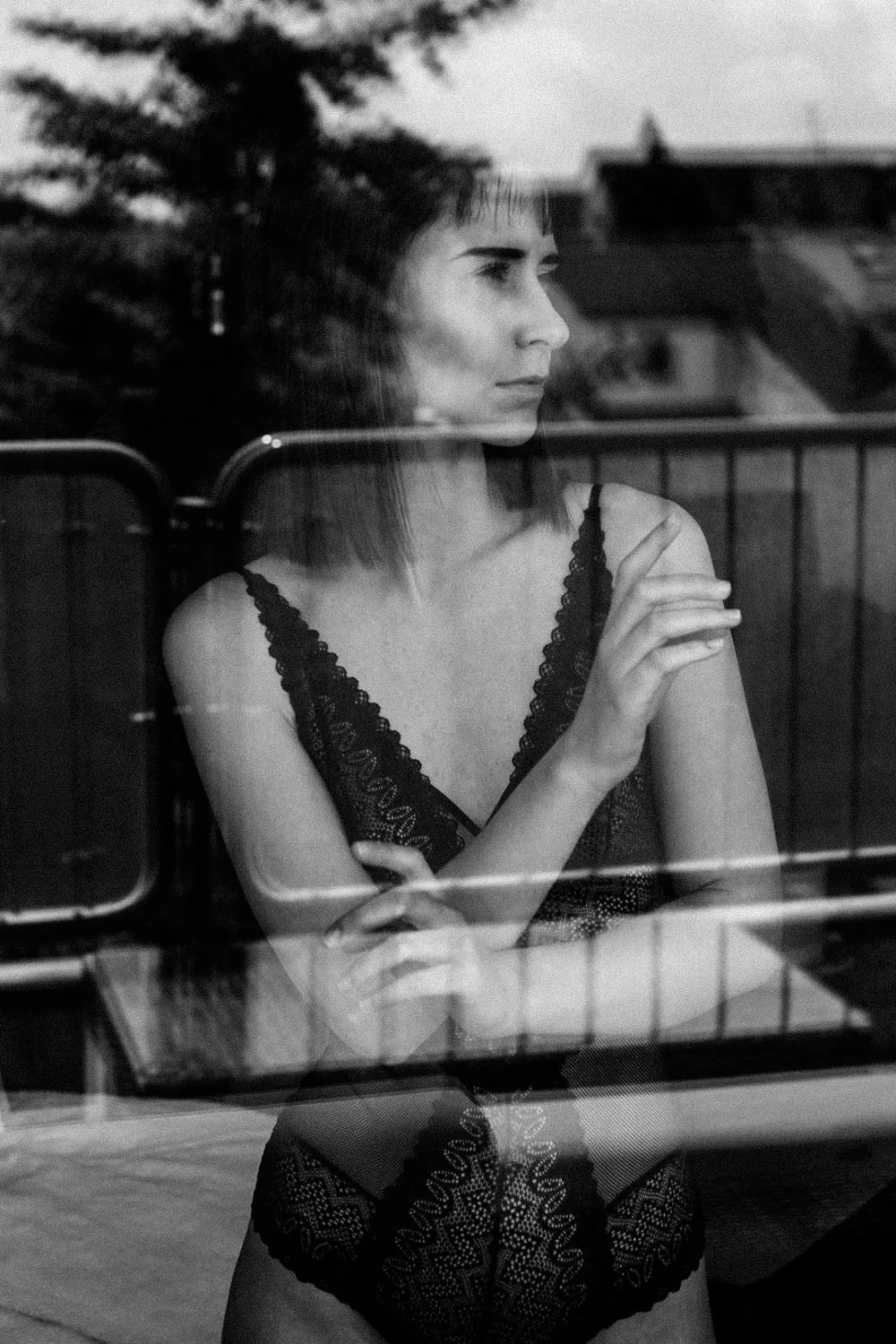 Schwarz-weiß-Fotografie eines nachdenklichen jungen Mädchens, das hinter einer Fensterscheibe steht, getragen in einem eleganten schwarzen Spitzenkleid.