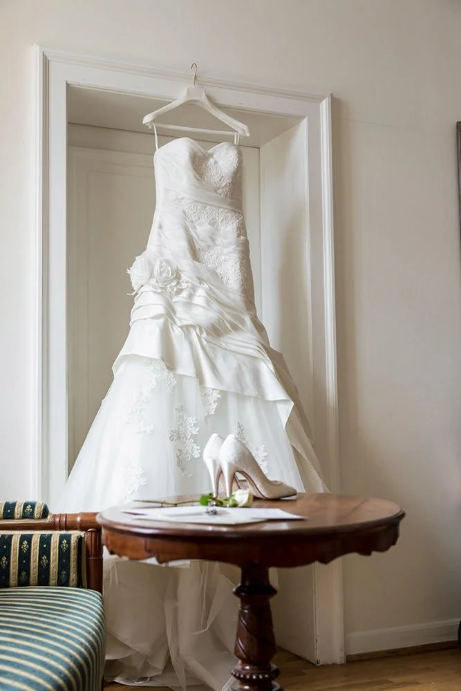 Ein weißes Brautkleid hängt an einem Haken in einer Türrahmen. Auf einem kleinen Holztisch im Vordergrund liegen weiße High Heels, eine Rose und eine Notiz.