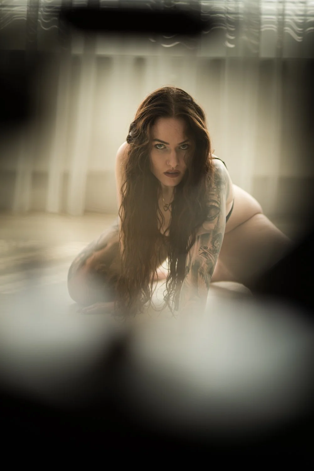 Junge Frau mit langen, dunklen Haaren, Tattoos, schaut intensiv in die Kamera, auf allen vieren auf einem Boden in einem hell erleuchteten Raum.