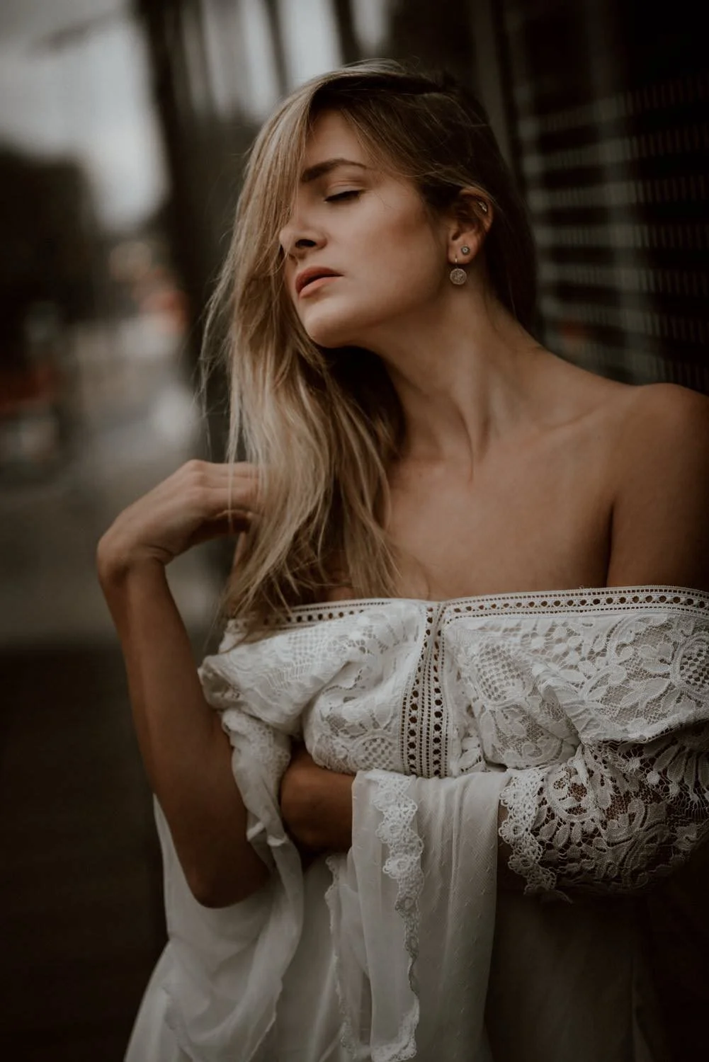 Eine Frau mit langen, gewellten, braunen Haaren, die eine weiße, lace-gefütterte Bluse trägt. Sie steht an einer dunklen Gitterwand, ihre Augen sind geschlossen, während sie ihre Hand am Hals hält.