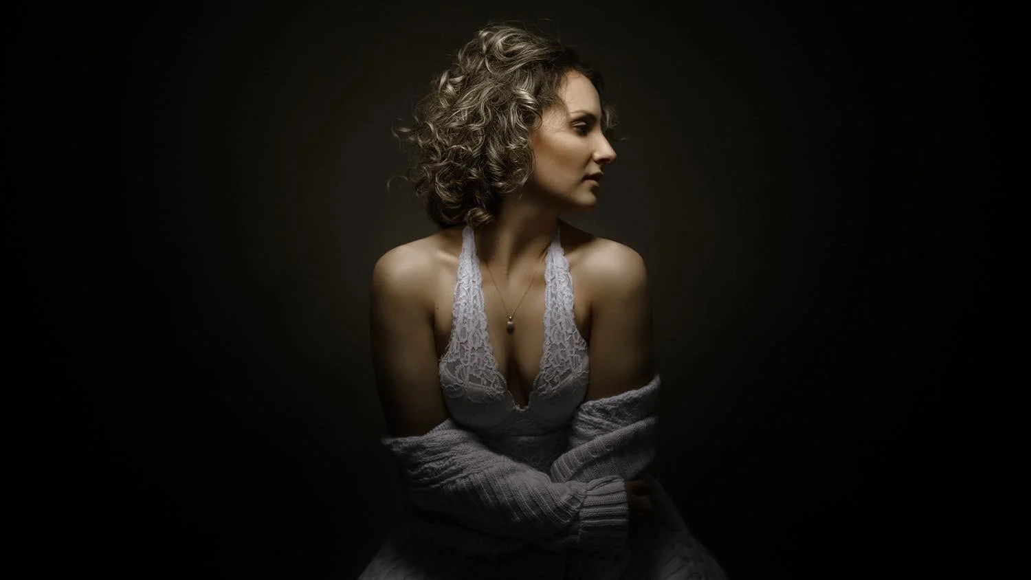 Eine junge Frau mit lockigem Haar in einem weißen Spitzenkleid und einer grauen Strickjacke vor dunklem Hintergrund, seitlich betrachtet.