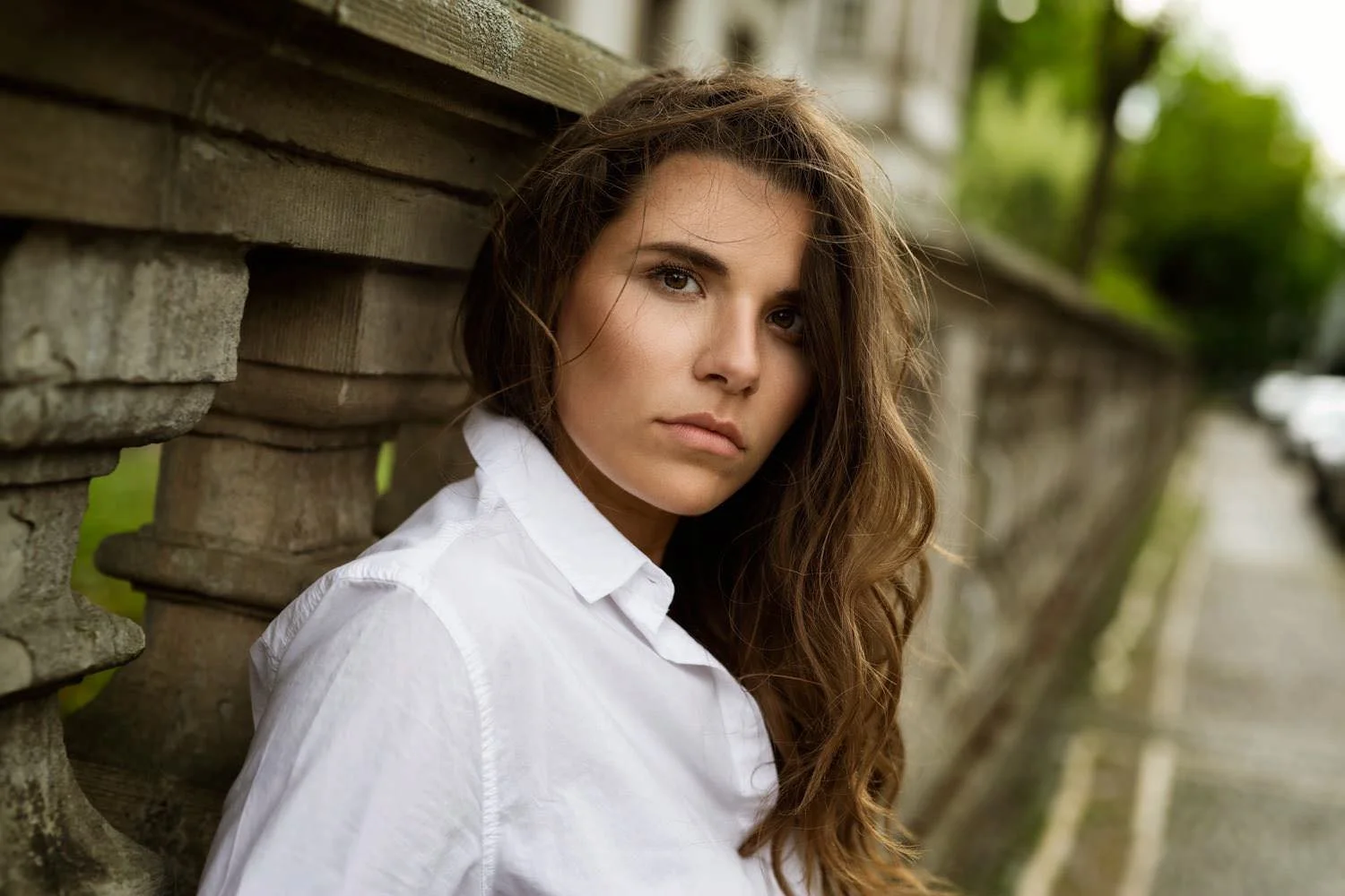Eine junge Frau mit braunen, lockigen Haaren, trägt ein weißes Hemd und lehnt an einem Holzzaun.