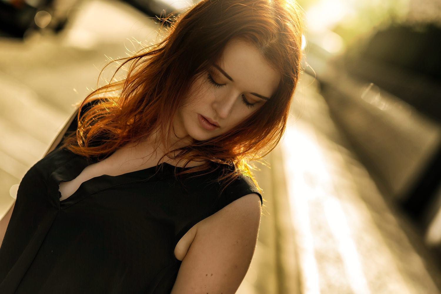 Junge Frau mit roten Haaren, in einem schwarzen Kleid, schaut nach unten, im Sonnenlicht, am Wasser. Hintergrund unscharf.