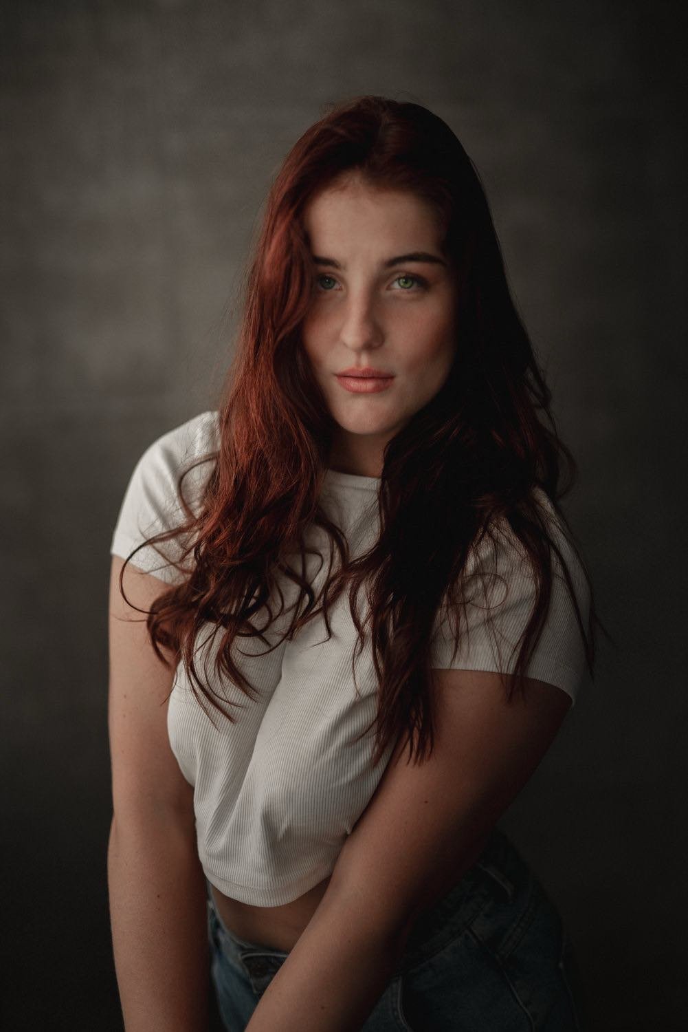 Junge Frau mit langen, lockigen braunen Haaren, trägt ein weißes T-Shirt und jeans, steht vor dunklem Hintergrund.