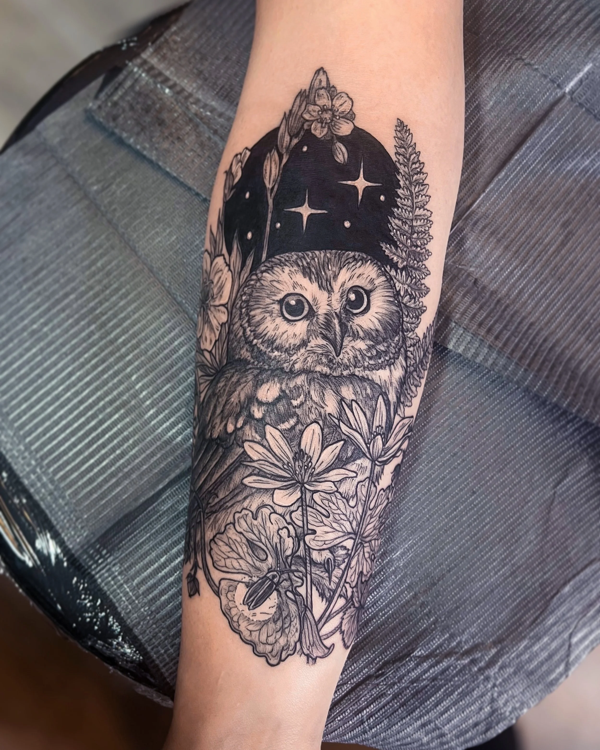 Forearm tattoo of a small owl with botanical elements and a firefly surrounding and a starry oval blackout night sky in the background