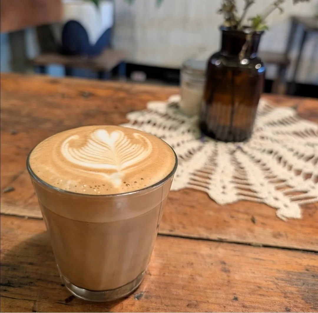 image of a coffee in a glass with decorative heart art on a wooden table, in the background is a doily and a plant in an amber glass vase