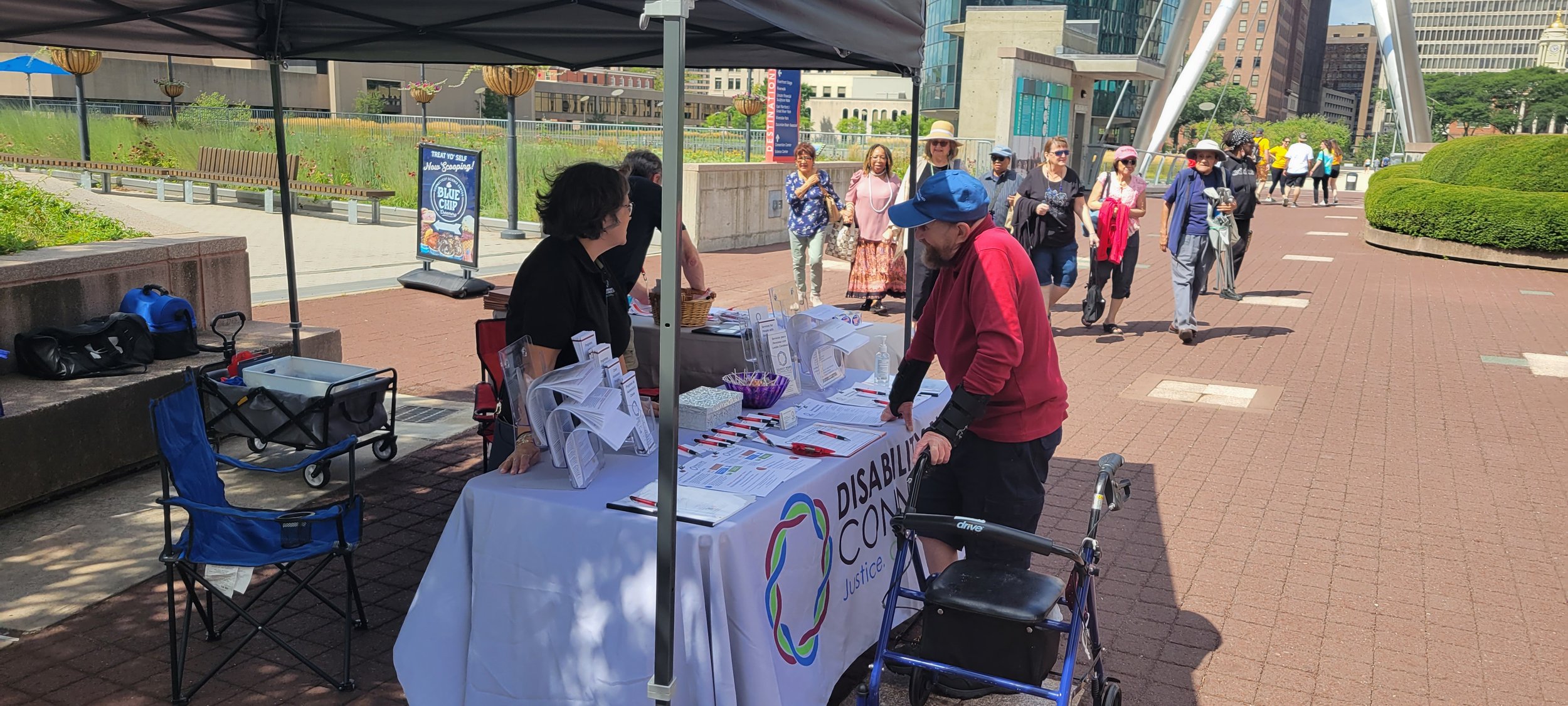  DRCT alum and volunteer Siobhan Morgan at the DRCT resource table talking with a man wearing a blue cap, red sweatshirt and black shorts, who is resting his right arm on the DRCT table, and his left hand is holding onto his walker that has wheels on