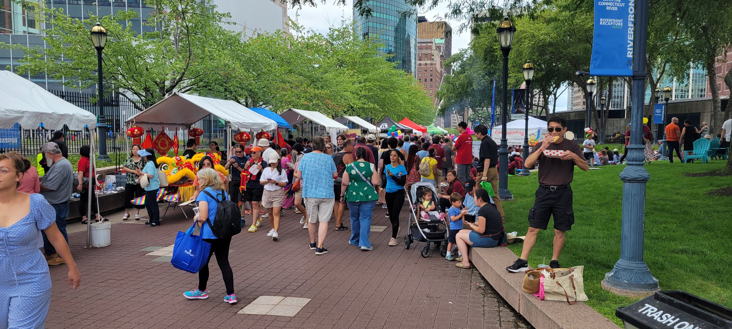  An event photo from the Riverfront Dragon Boat + Asian Festival that shows nine vendor tents set up on the left side of the Mortensen Riverfront Plaza and one face painting vendor tent on the right side on a cloudy day. People can be seen walking al