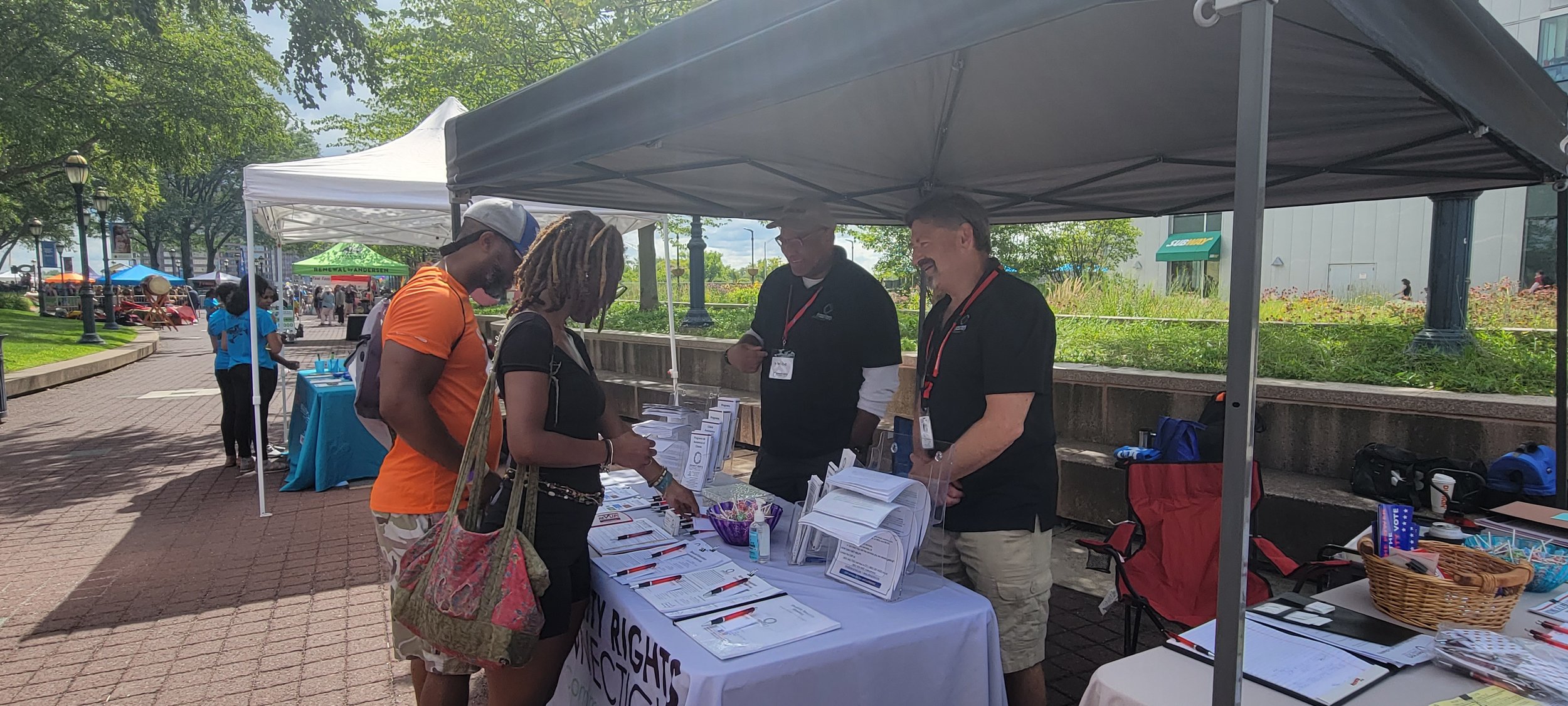 DRCT’s Strategic Partnerships Manager, Mike Whilby and Lead Investigator, Jim Welsh stand smiling under the DRCT black tent while talking to two event attendees at the DRCT resource table. 