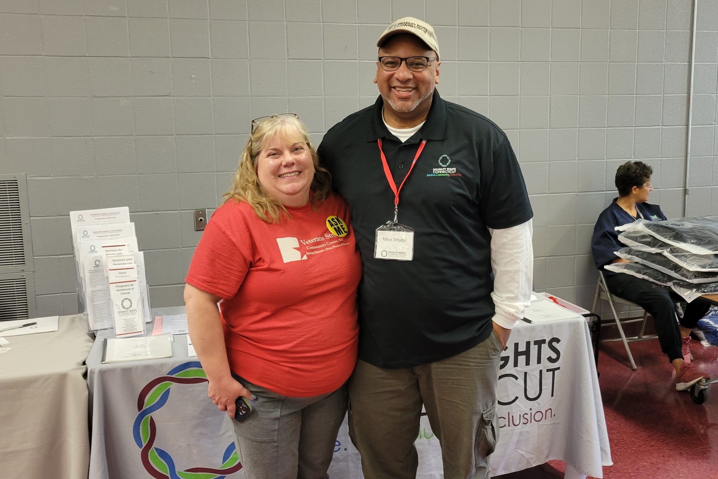   DRCT’s Strategic Partnerships Manager, Mike Whilby stands smiling (on the right side) with Donna M. Dognin, Co-Founder/Executive Director of Veterans Strong Community Center, Inc in front of DRCT resource table with two individuals sitting at a tab