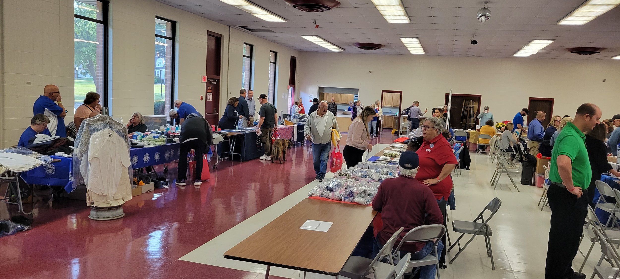   Group of individuals standing and sitting at several rows of vendor tables while talking and checking out program materials, clothing items and household supplies.  