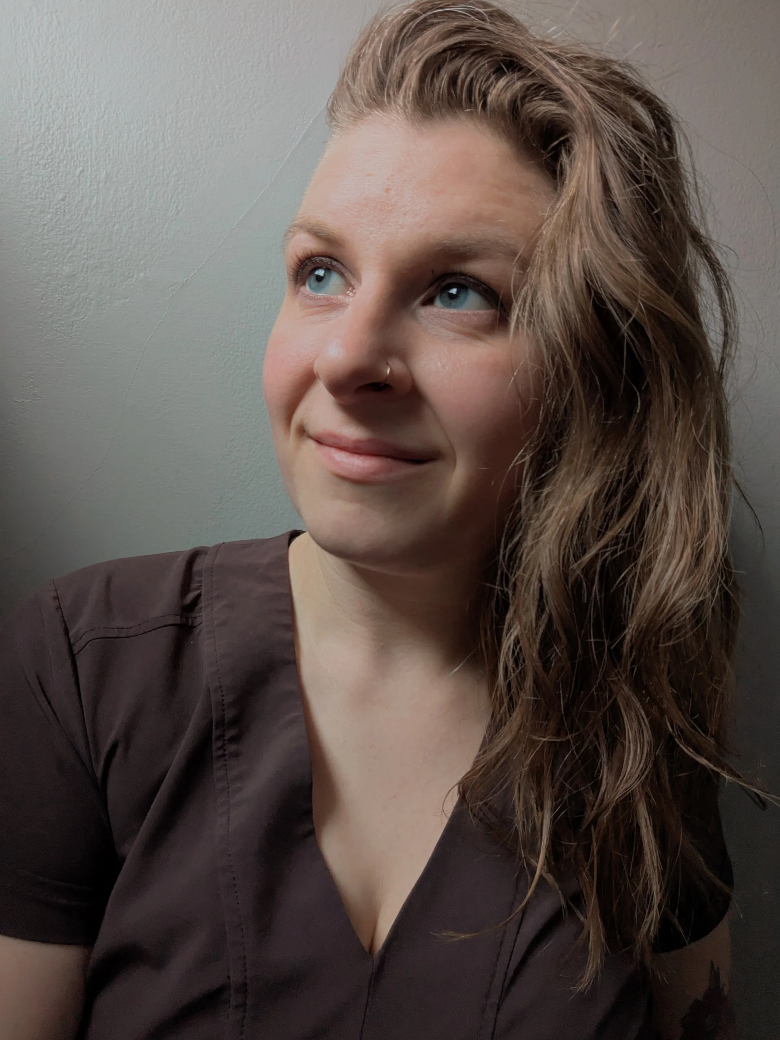 A light-skinned person with light brown hair and blue eyes looks off camera. They are wearing a brown shirt, and stand against a white wall.