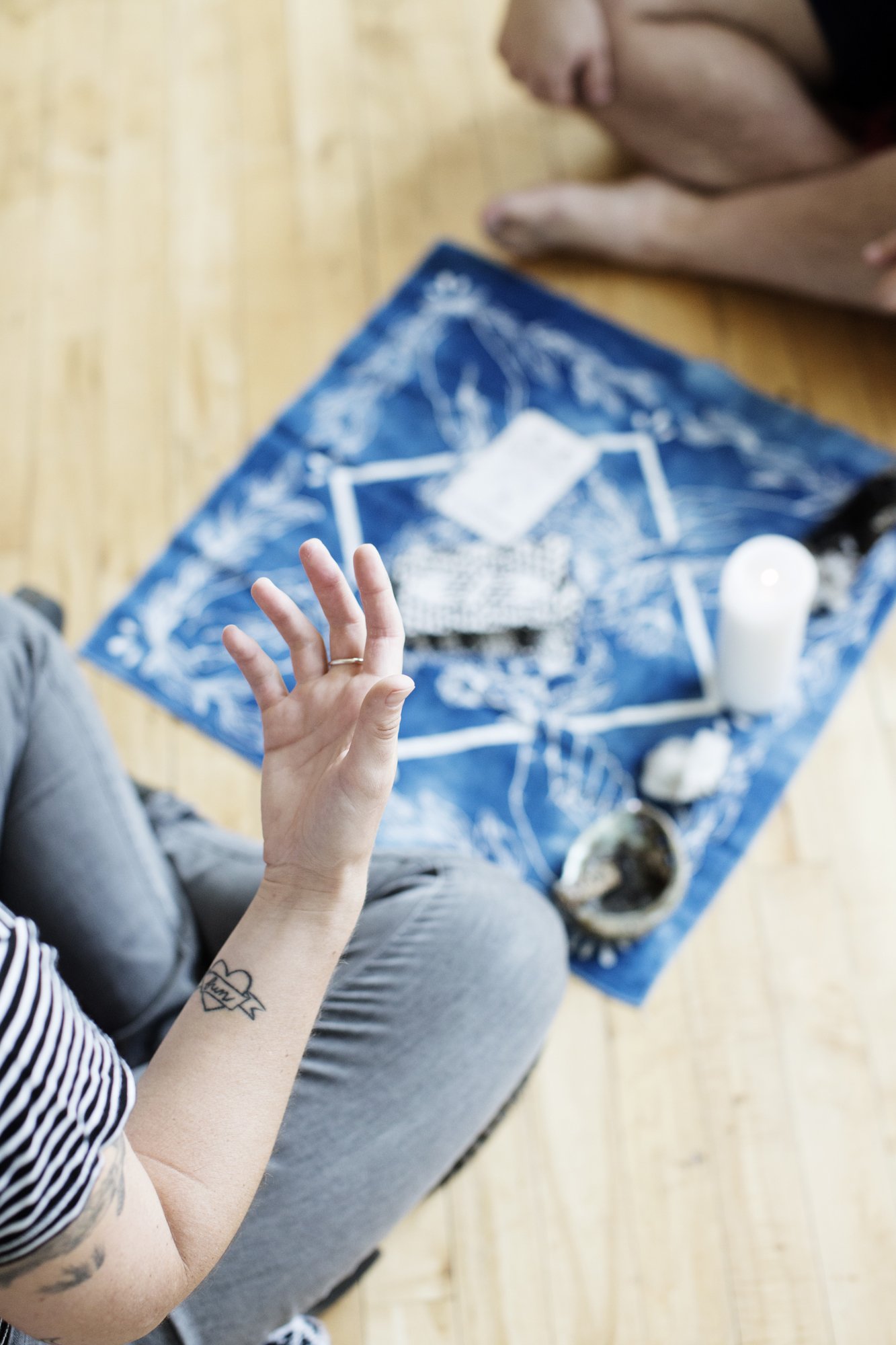 a light skinned person with tattoos sits crossed legged on the floor in front of a client with a tarot spread laid out between them