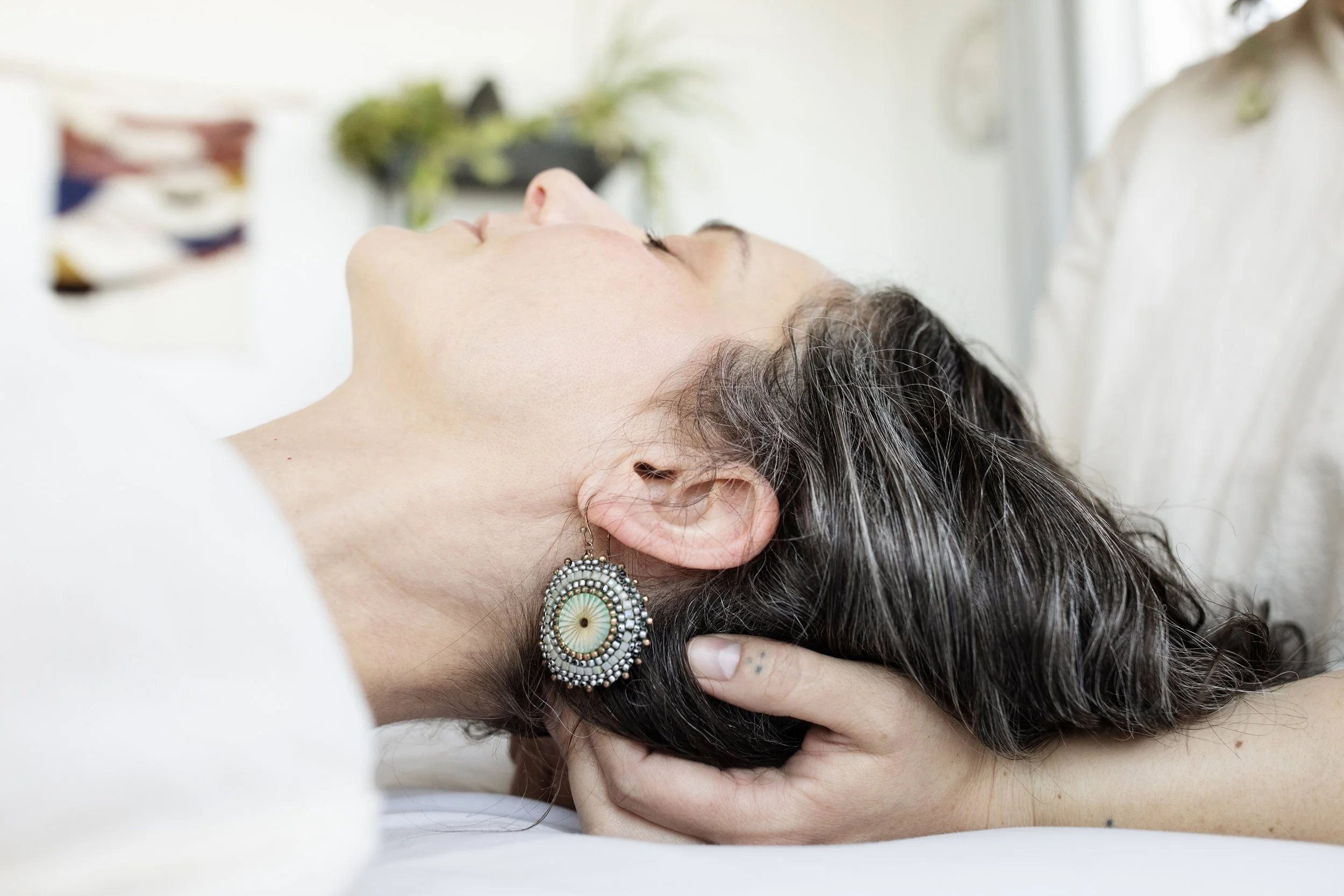 A female patient with light skin and dark hair reclines on a massage table. A practitioner with light skin holds her head, applying gentle pressure to her neck.