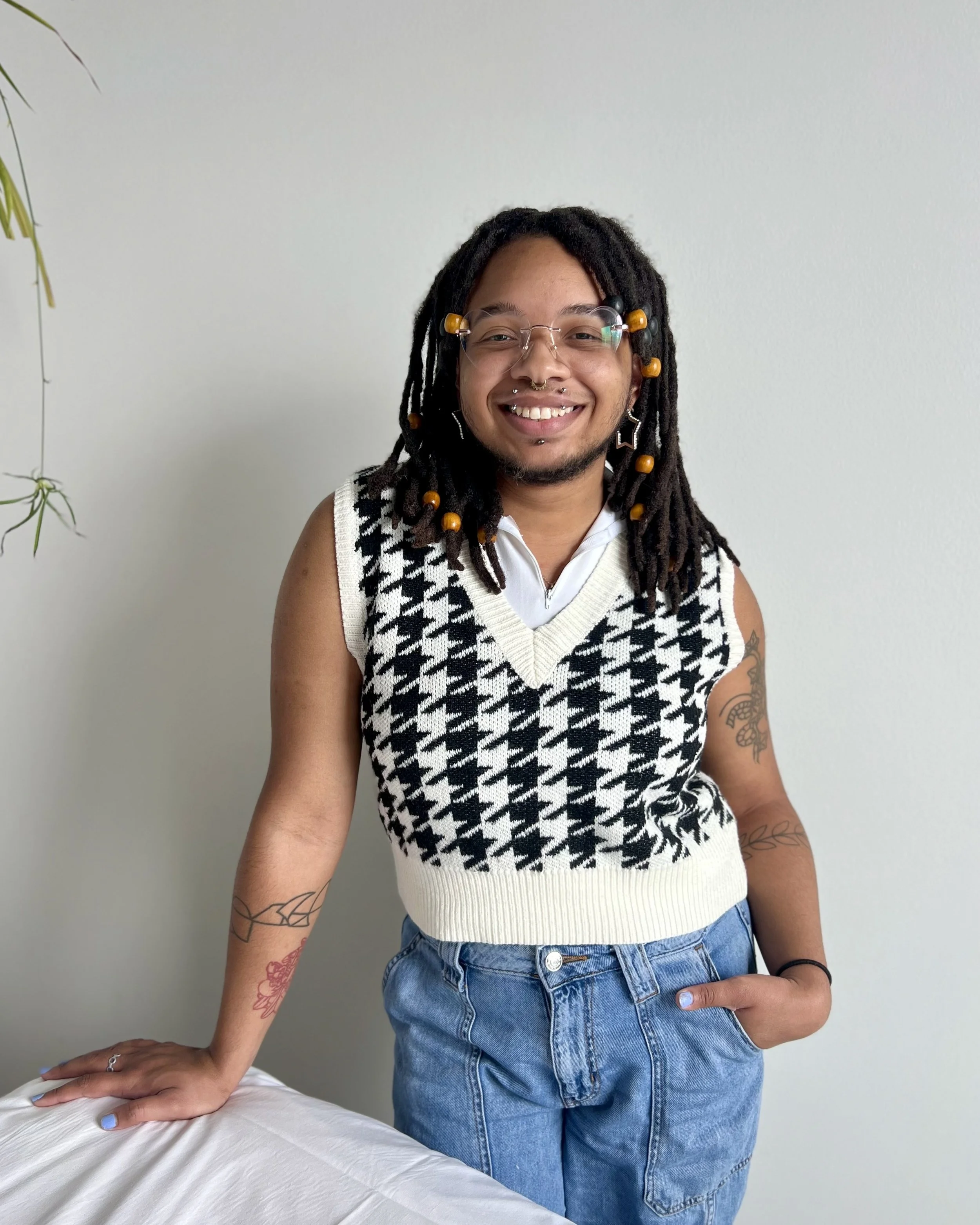 A person with medium-dark skin and dark hair stands against a white wall in a treatment room. He is smiling at the camera. He is wearing heart-shaped glasses, a checkered vest, and jeans.