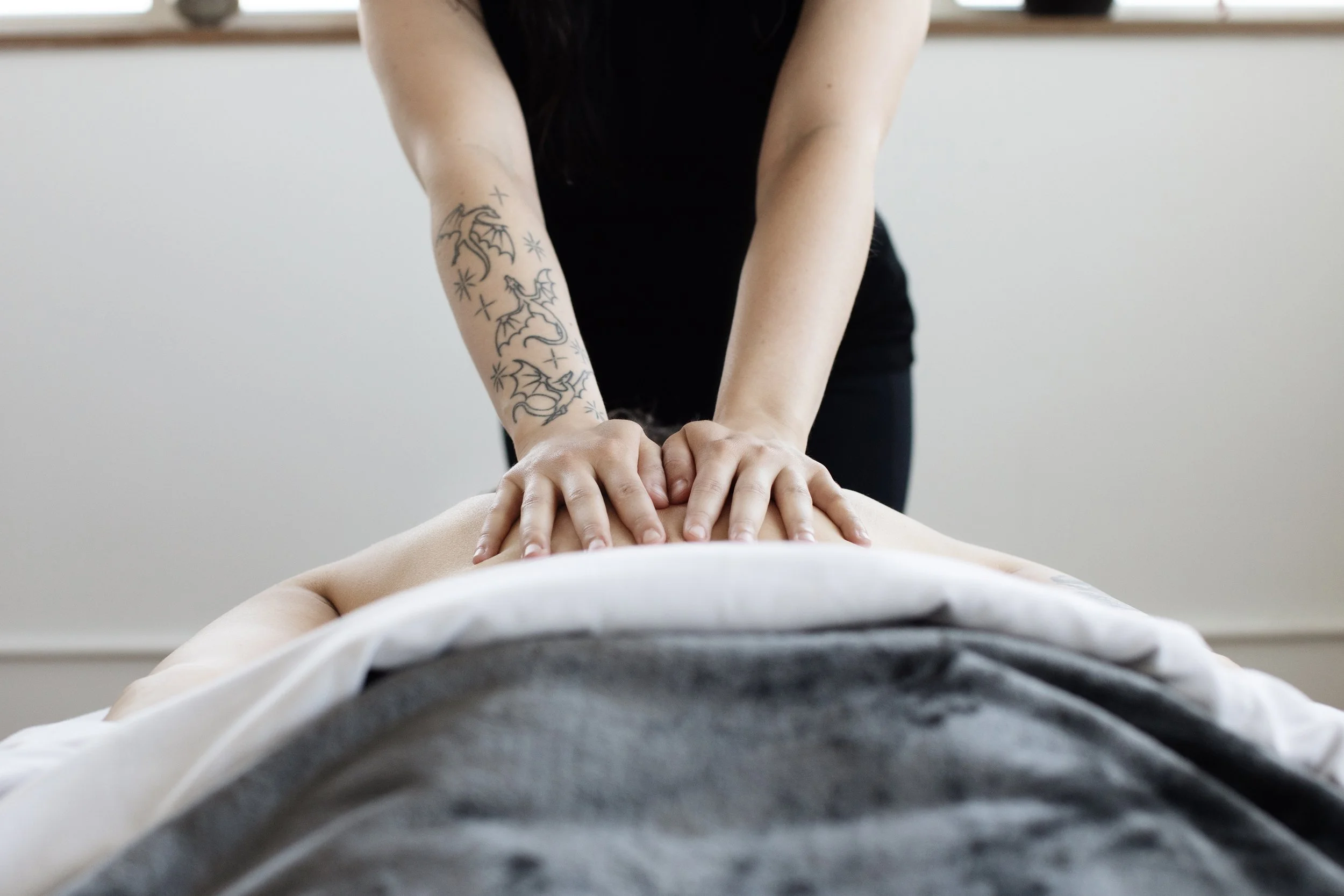 A patient with light skin lies face down on a massage table, and receives massage from a practitioner with light skin and tattoos.