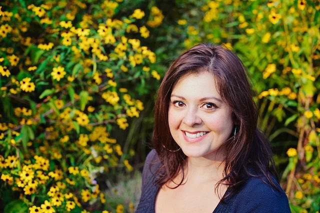 A woman with long brown hair, brown eyes, and medium skin, and a navy shirt smiles warmly at the camera, standing in front of yellow flowers.