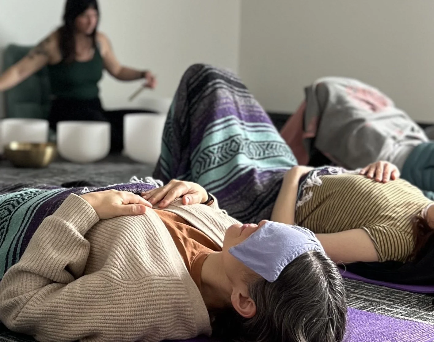 Three people are reclining on yoga mats, with blankets and eye pillows, listening to a sound bath. A sound healing practitioner is in the background, playing crystal singing bowls.