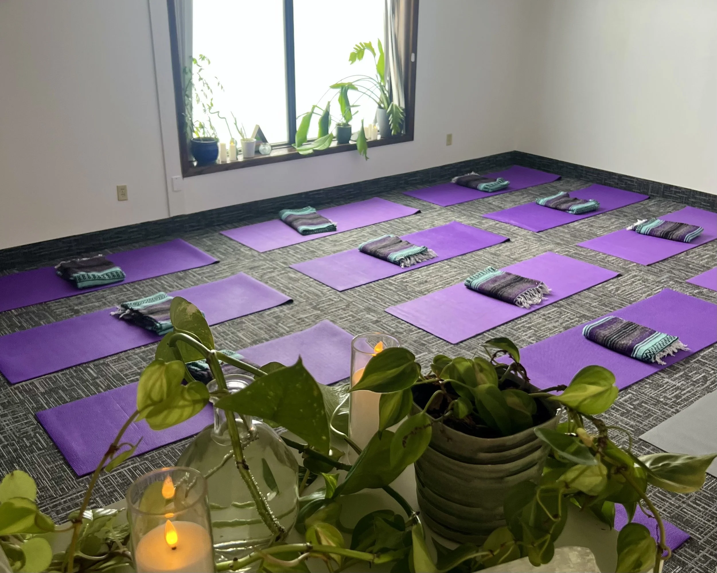 Purple yoga mats and striped blankets are spread on the floor of a space. Plants are in the foreground, and in the background, a window with plants and candles on the windowsill allows a gentle light into the space.