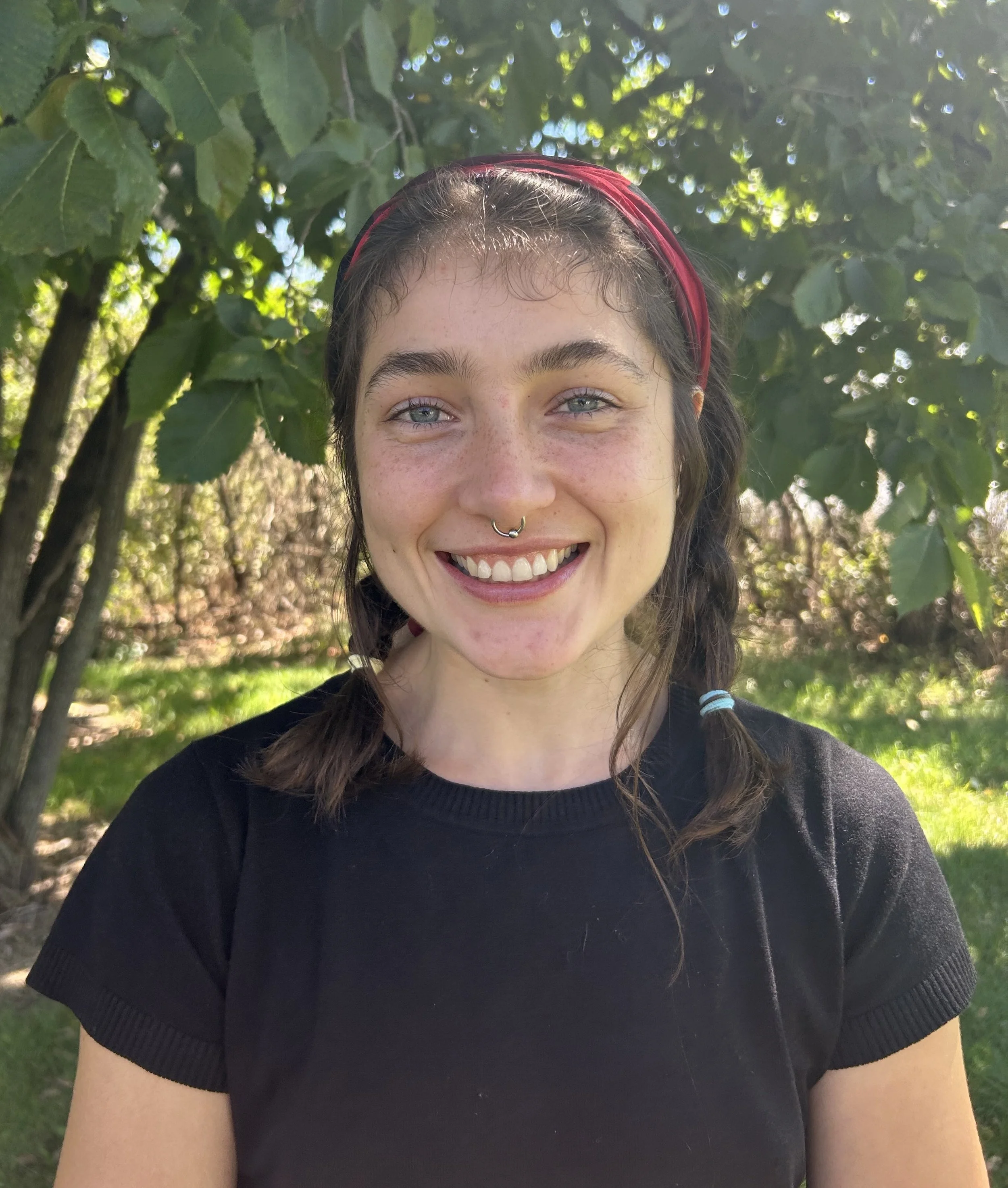A light skin person with dark brown hair in braids and a black tshirt stands in front of a leafy, green background.