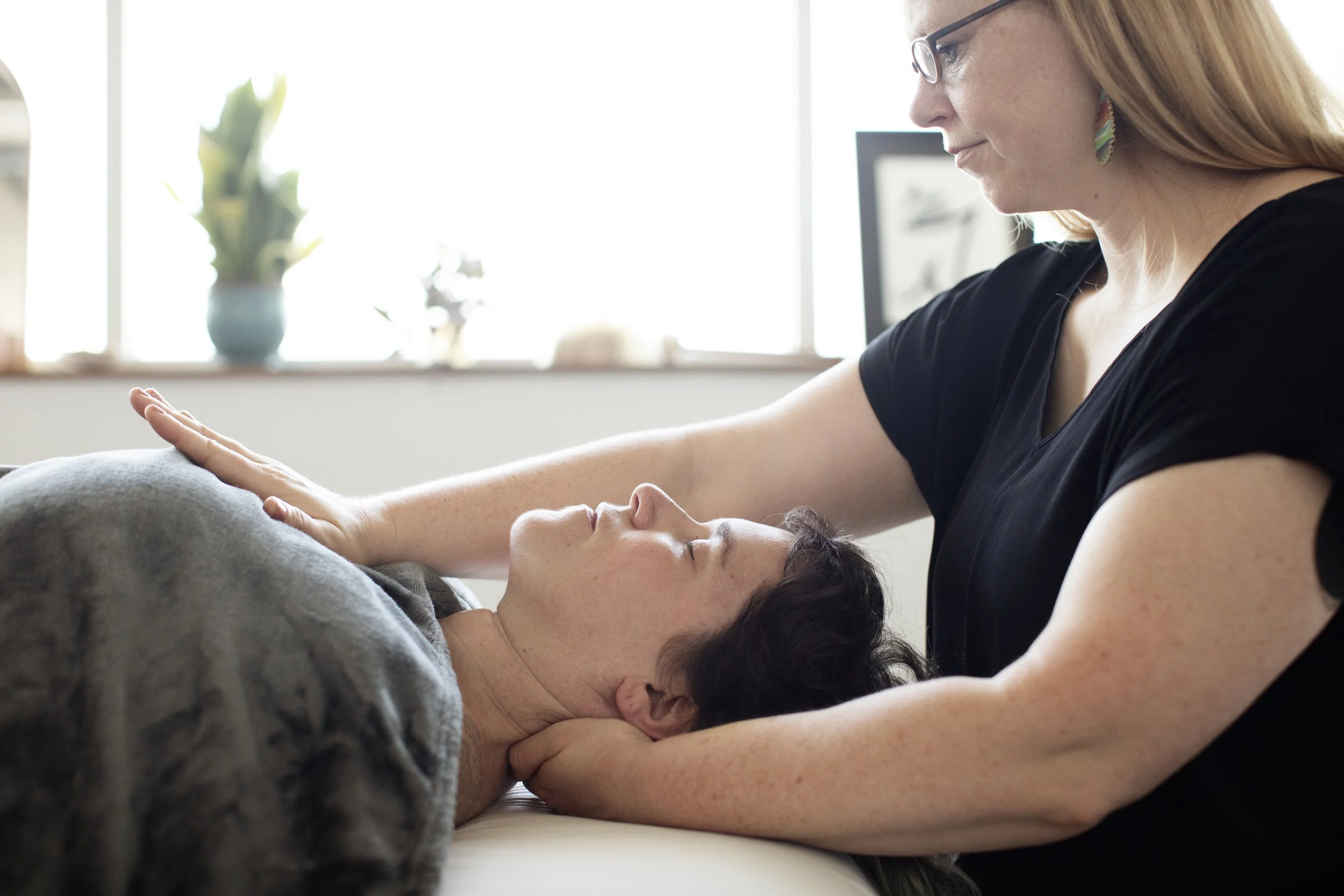 A practitioner with light skin holds the neck and chest of a light-skinned patient who is resting on the massage table under a blanket.