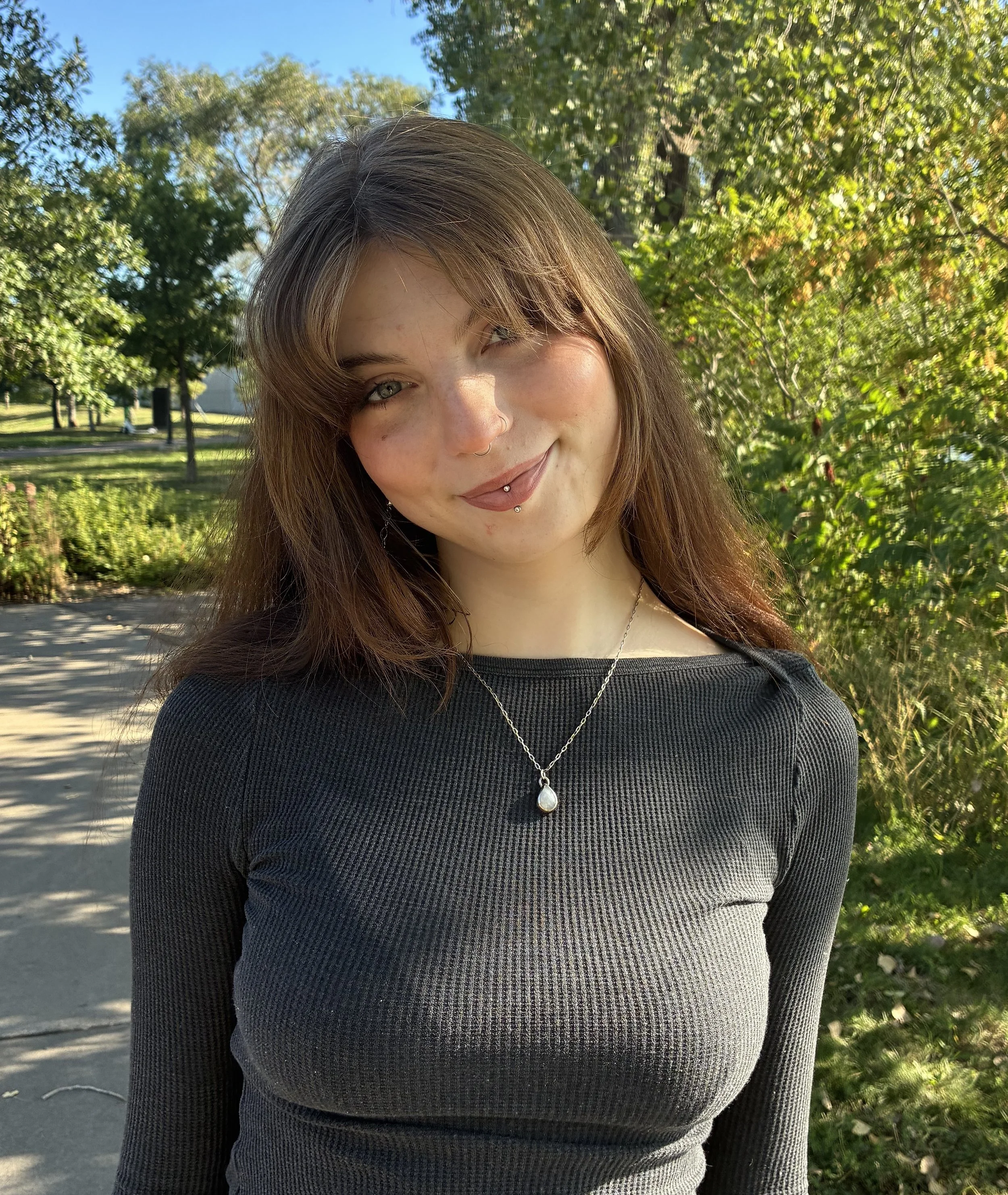 A woman with light colored skin and brown hair is smiling, while she stands in front of a beautiful green area with grass and trees.