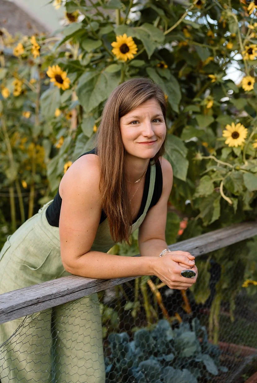 a woman with light skin and long brown hair leans against a fence, wearing overalls and a tank top. she stands in front of sunflowers.